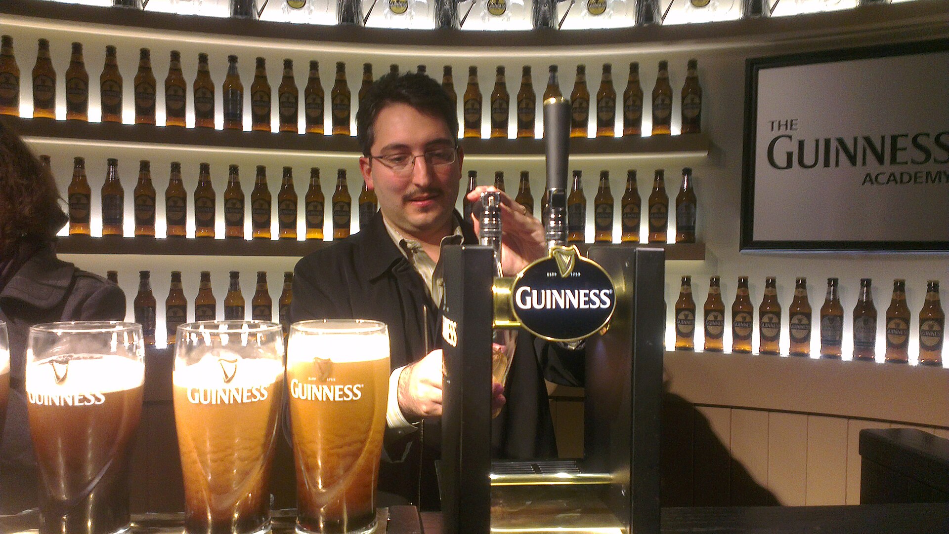 An adult visitor pours a pint at the Guinness Academy inside Dublin’s Guinness Storehouse, with filled glasses on the counter.