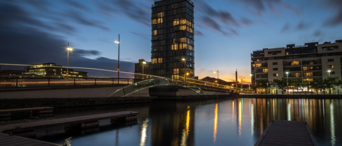 Alto Vetro apartment tower beside Dublin's Grand Canal Dock at dusk, with illuminated windows and bridge lights reflected in the water.