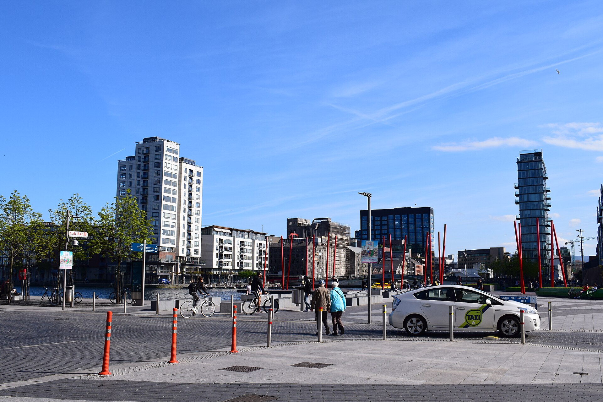 Apartment buildings surround Grand Canal Dock in Dublin, with cyclists, pedestrians and a taxi crossing the paved square in sunshine.
