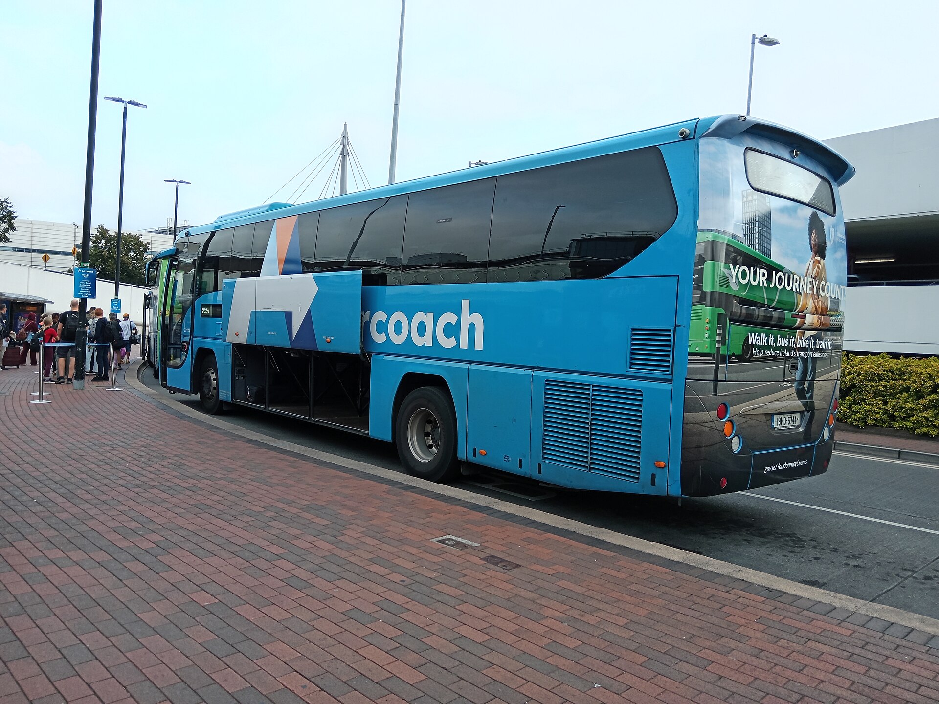 A blue Aircoach waits at Dublin Airport with its luggage compartment open and travellers queuing beside the pavement.