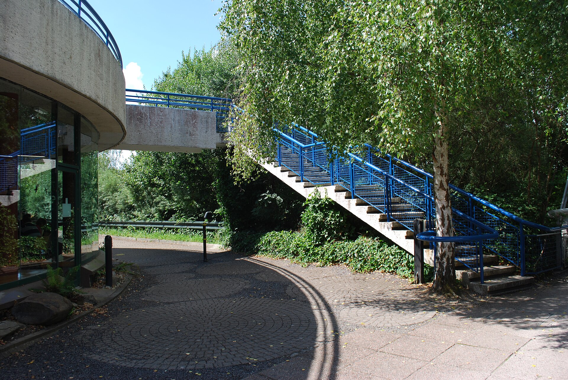 Blue-railed steps climb beside the curved exterior of the Bru na Boinne Visitor Centre, surrounded by leafy trees.