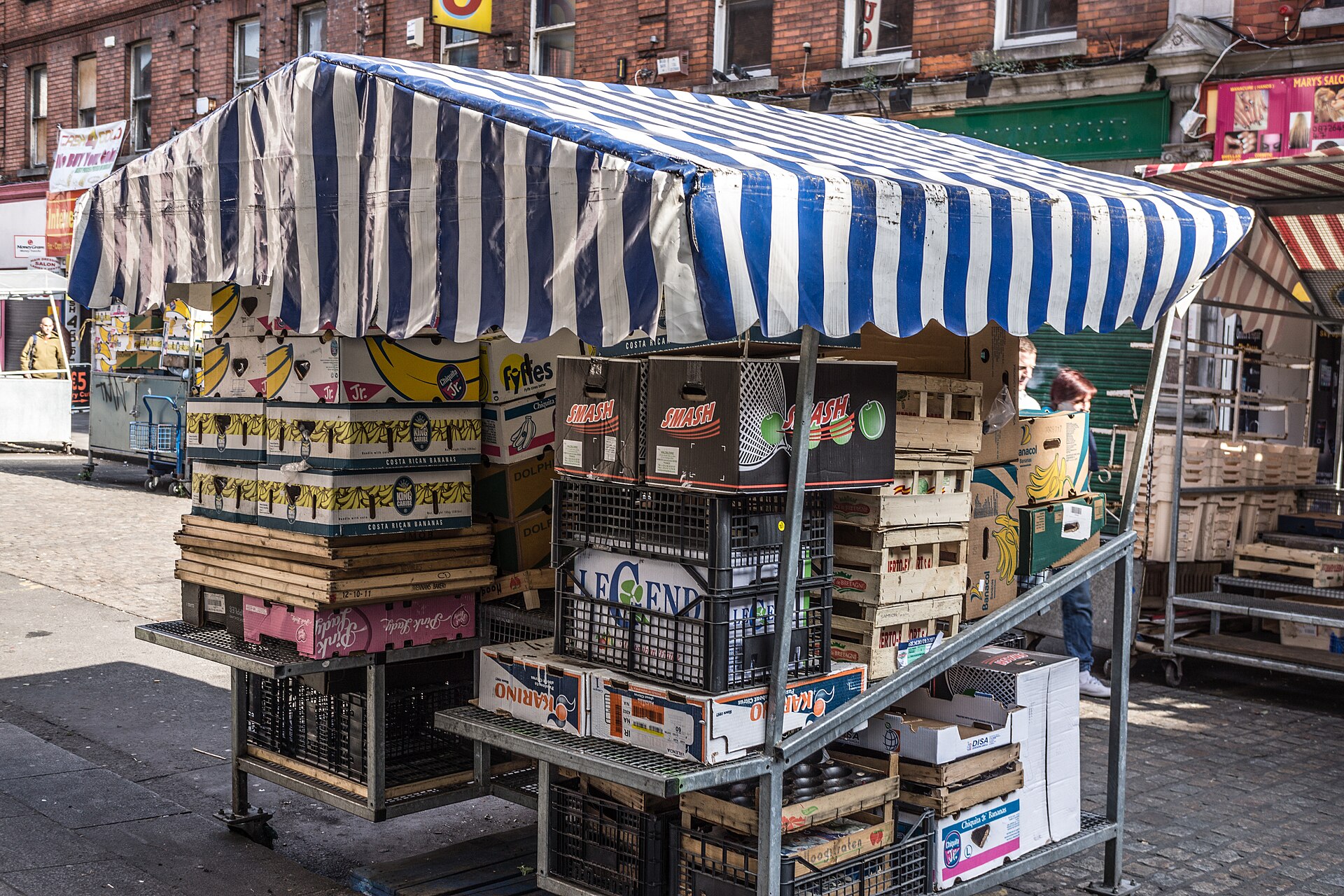 A blue-and-white striped canopy covers stacked crates and boxes at a Moore Street market stall in Dublin.