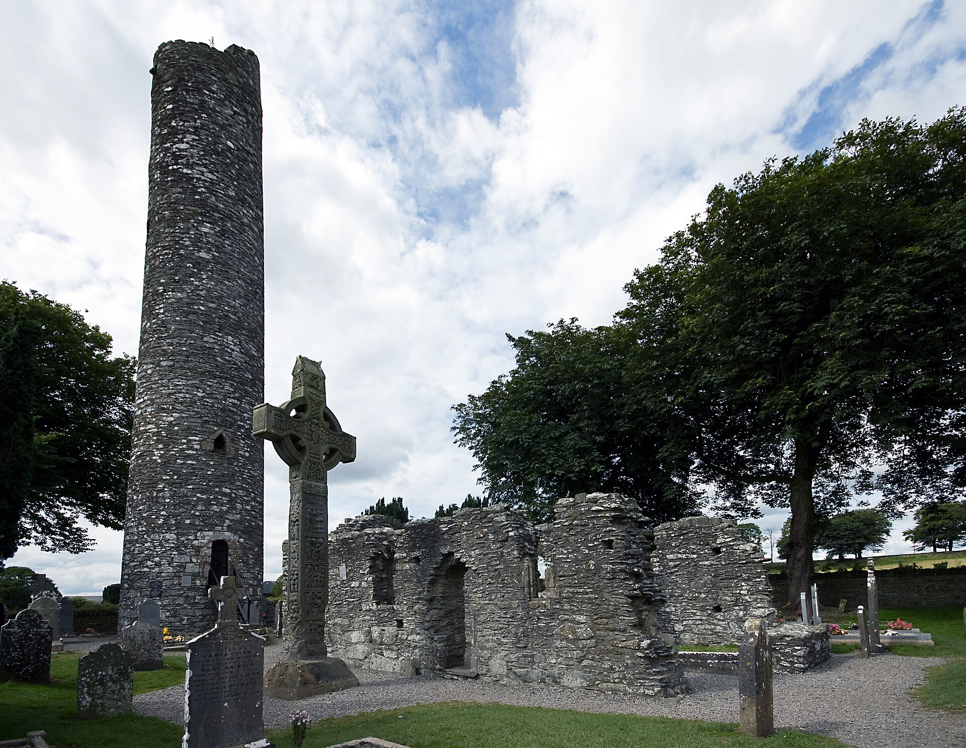 A carved high cross stands beside the round tower and ruined church walls at Monasterboice, surrounded by gravestones and trees.