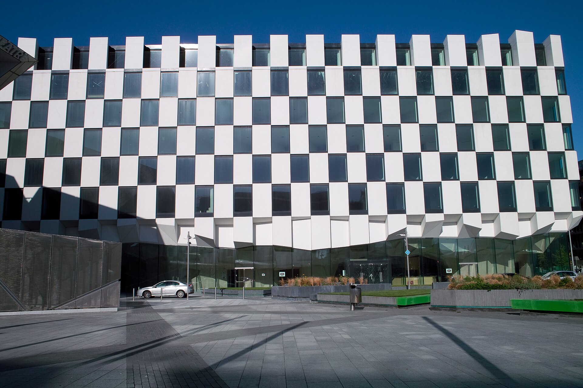 Chequered black-and-white facade of The Marker hotel rising above the paved Grand Canal Square in Dublin Docklands under a clear blue sky