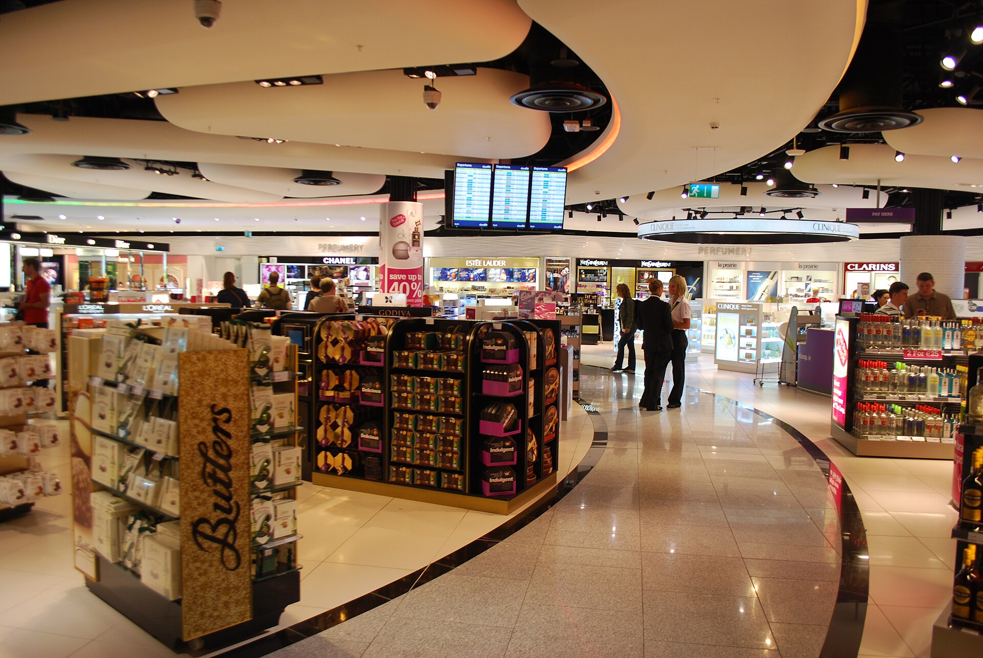 Chocolate and perfume displays fill the curved shopping hall at Dublin Airport Terminal 2, with travellers browsing between counters.