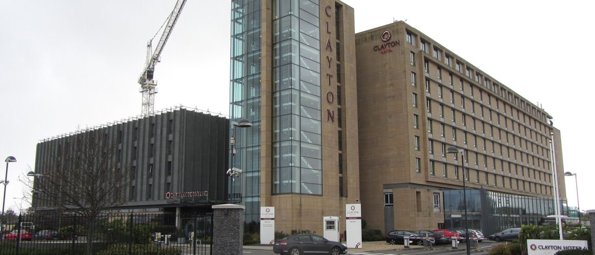 Clayton Hotel Dublin Airport, with its glass stairwell tower, entrance driveway and parked cars beneath an overcast sky.