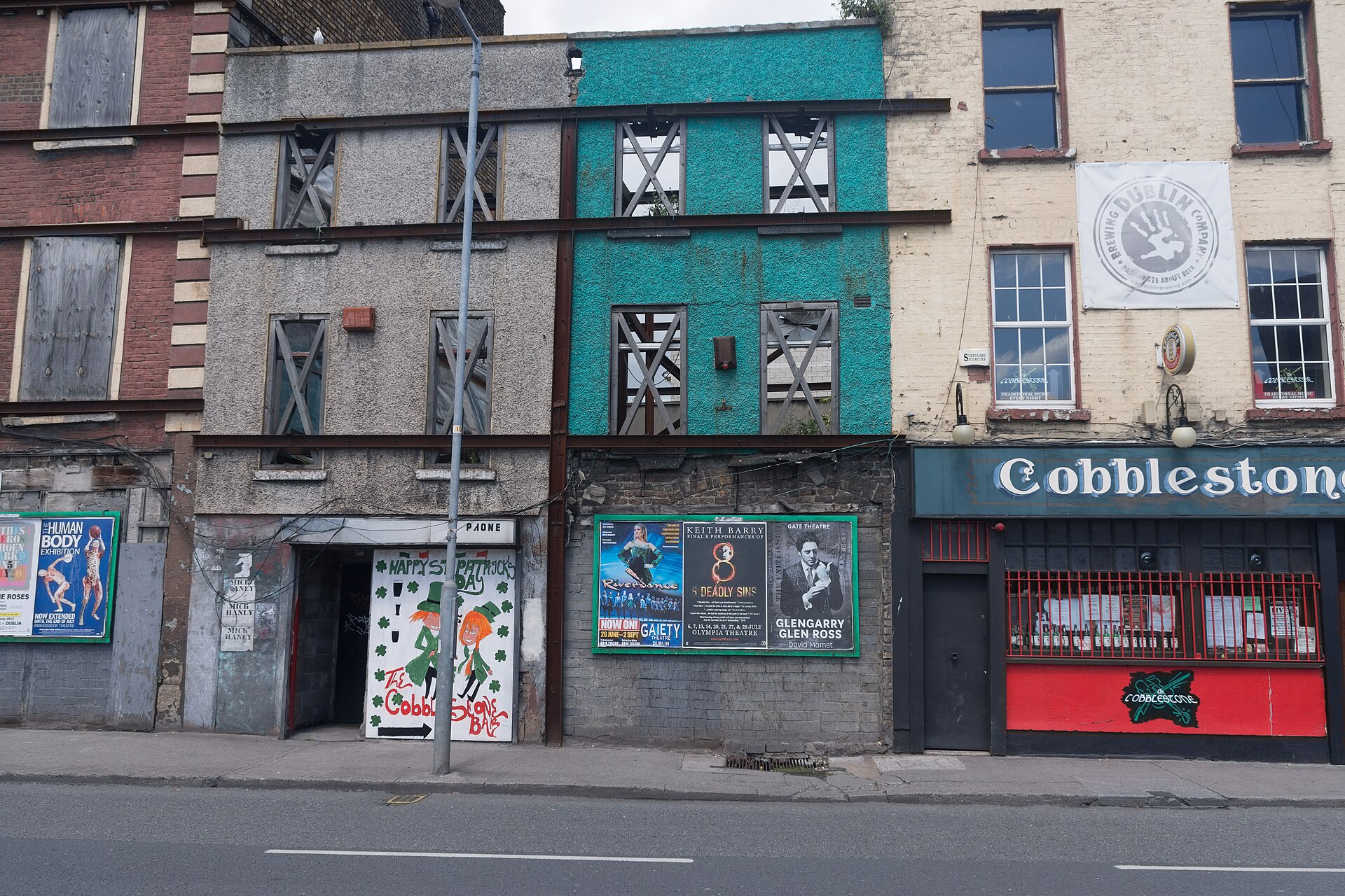 The Cobblestone pub in Dublin beside painted facades, with a red barred window and a brewing banner overhead.