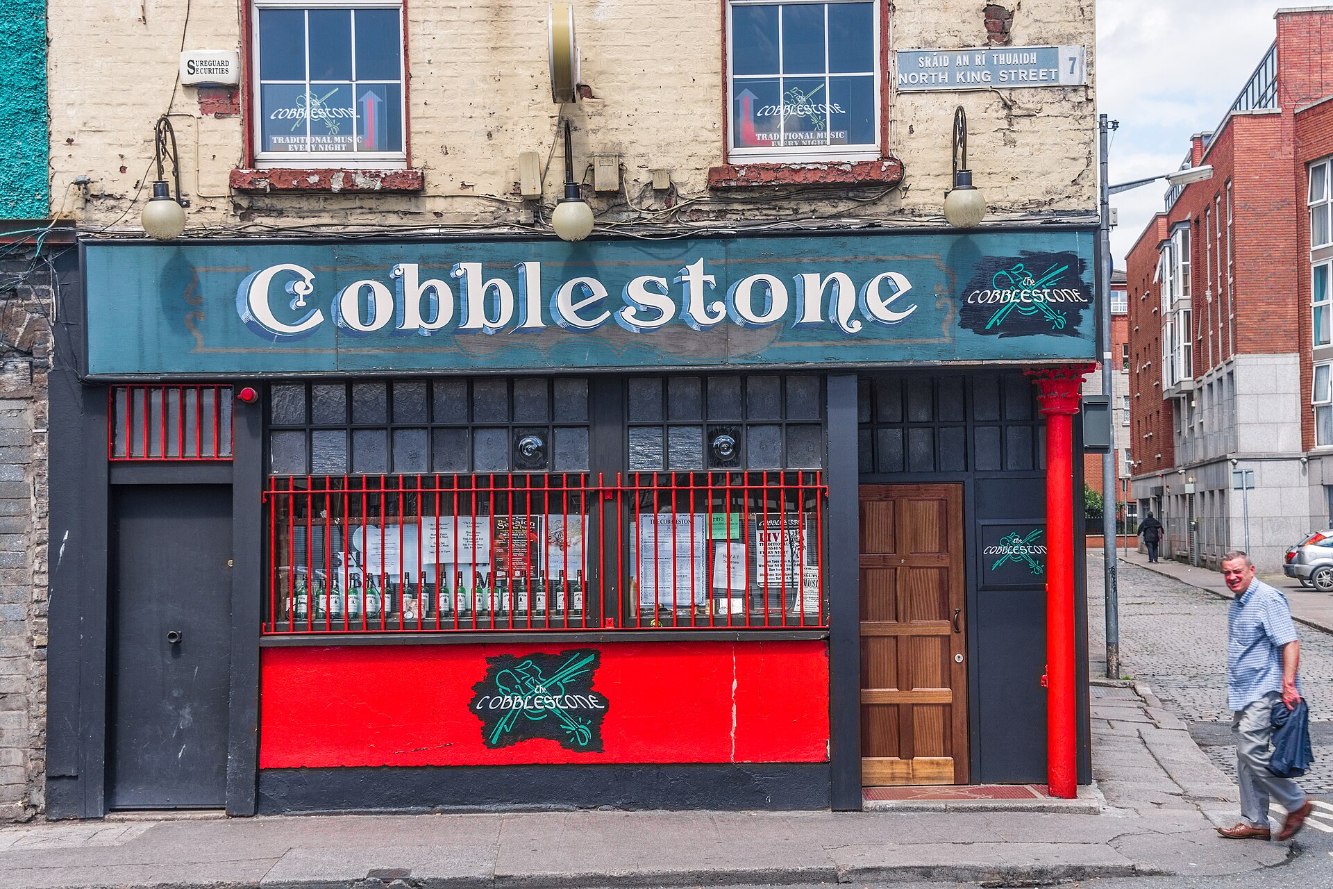 The Cobblestone pub entrance in Dublin, with a large blue sign, red window bars and a wooden door.