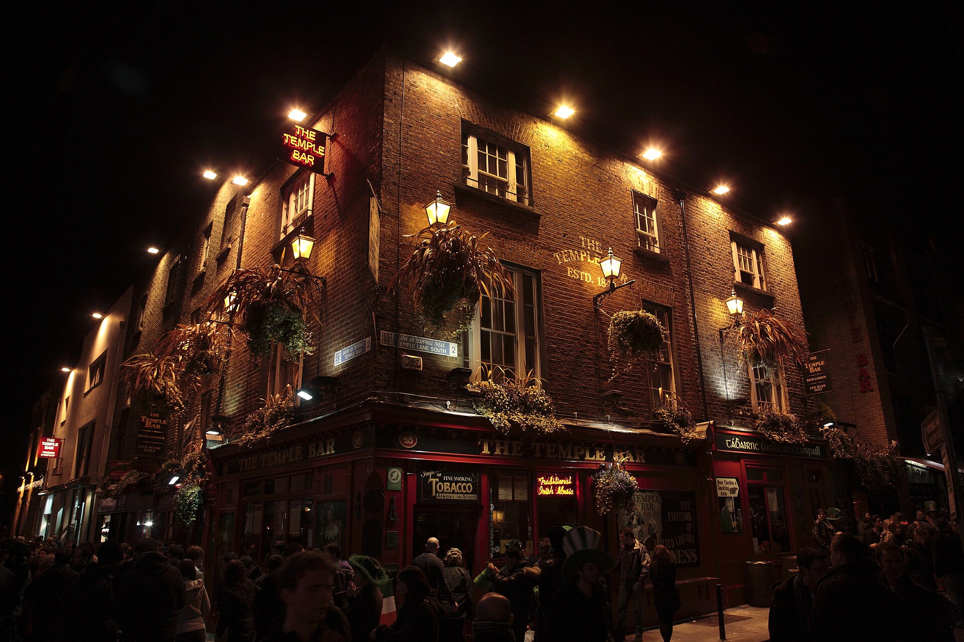 A crowd stands outside the illuminated Temple Bar pub in Dublin, beneath hanging baskets and a row of glowing lamps.