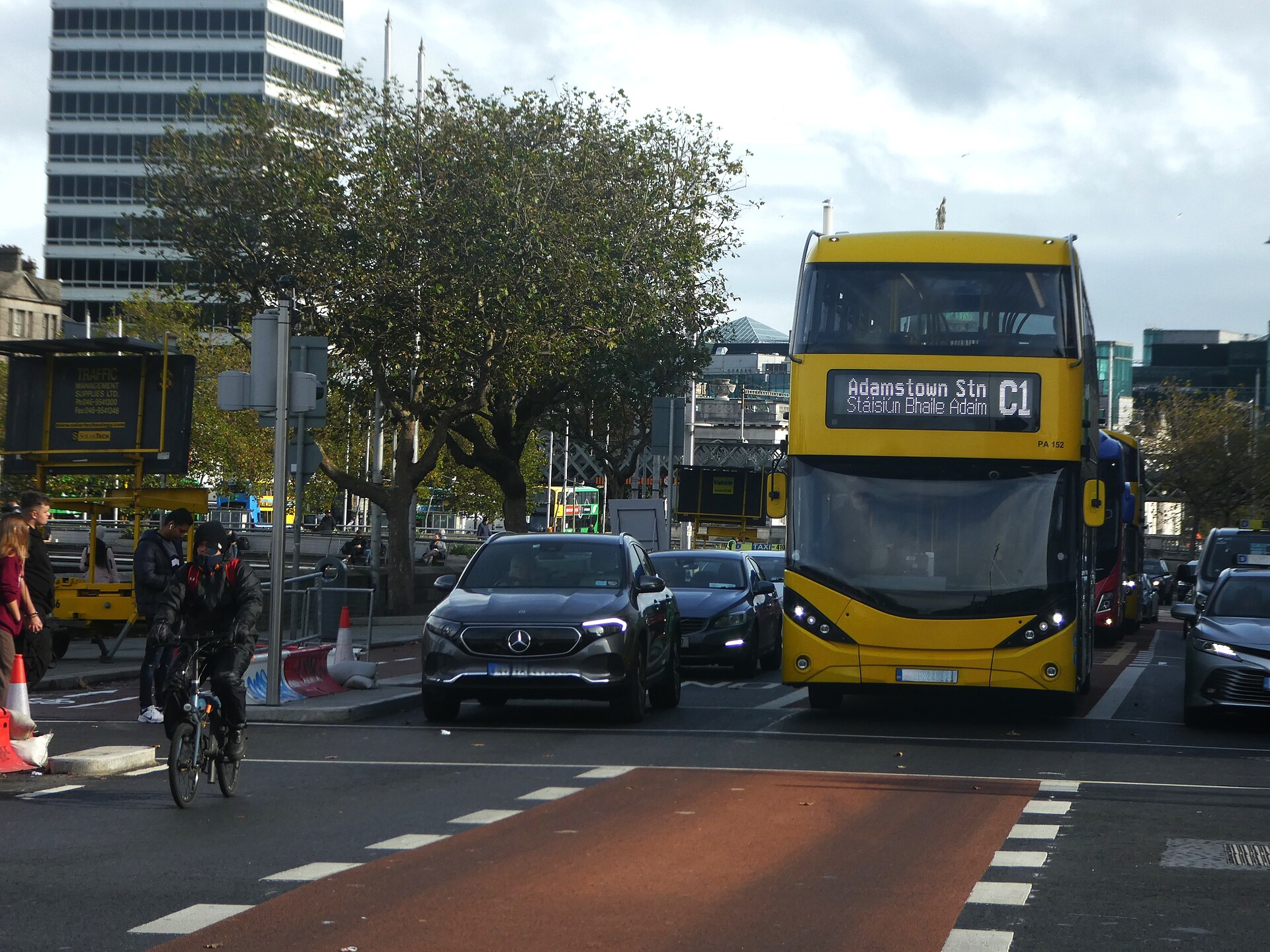 A cyclist rides beside cars and yellow double-decker buses on a busy roadway on Dublin's quays in daylight.