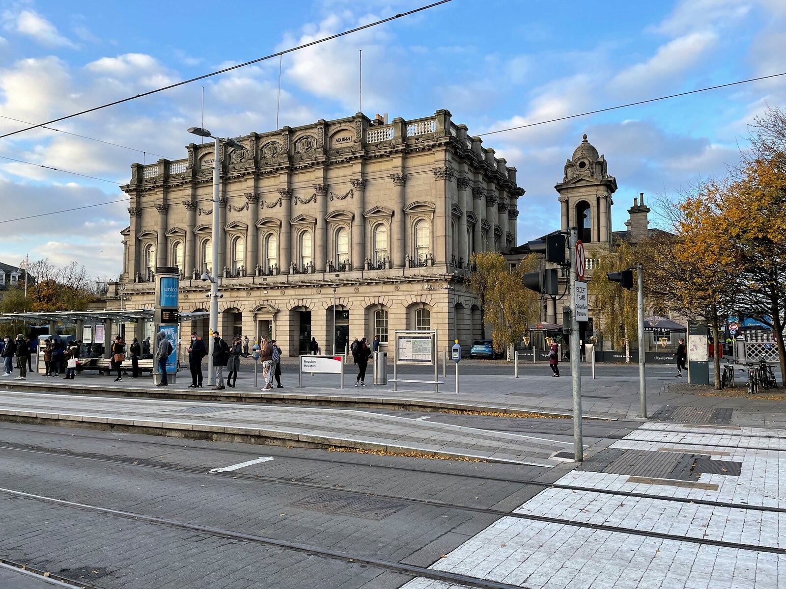 Dublin Heuston station's ornate stone facade overlooks tram tracks and people waiting on the forecourt in autumn sunshine.