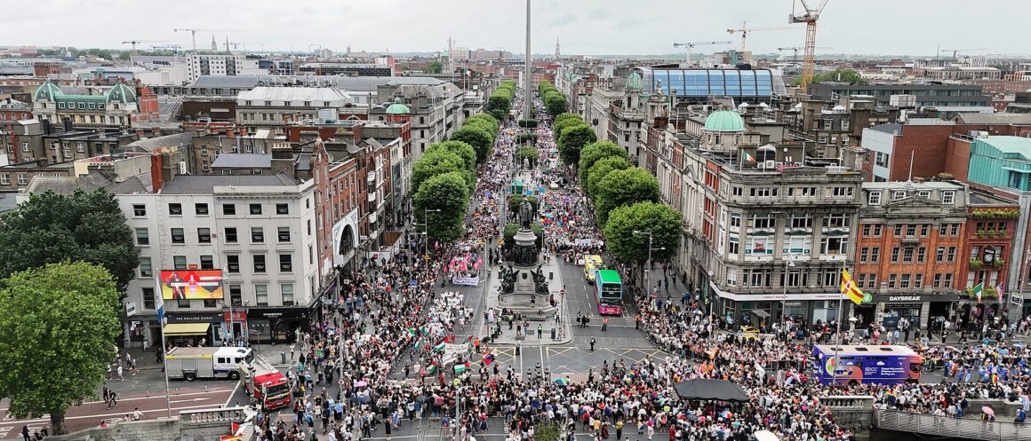 Dublin Pride crowds fill O’Connell Bridge and O’Connell Street, with rainbow flags below the Spire and O’Connell Monument.