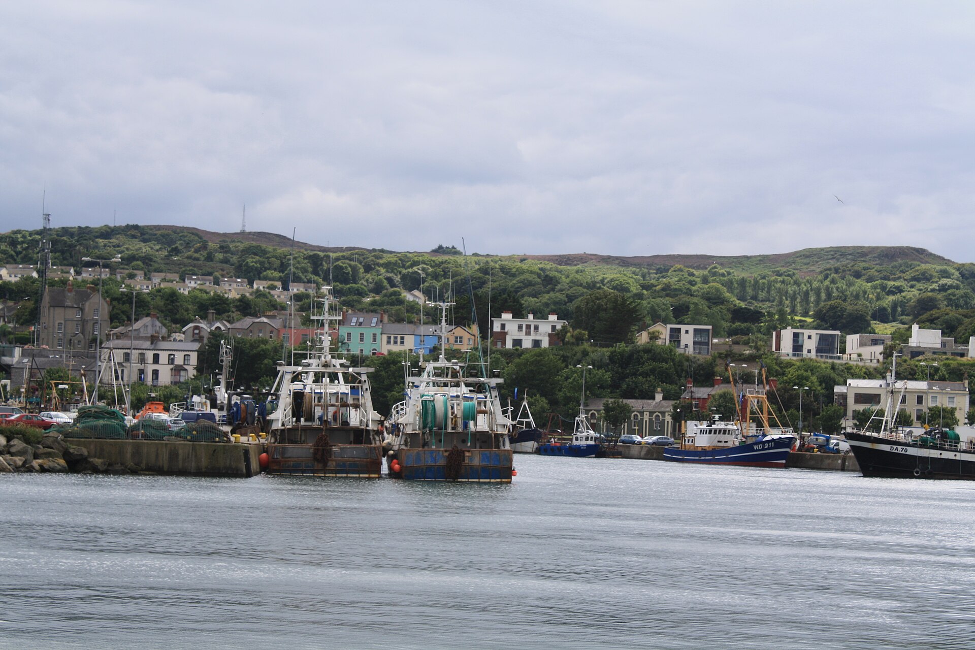 Fishing boats lie along the quays in Howth Harbour, below village houses and a green hillside under a cloudy sky.
