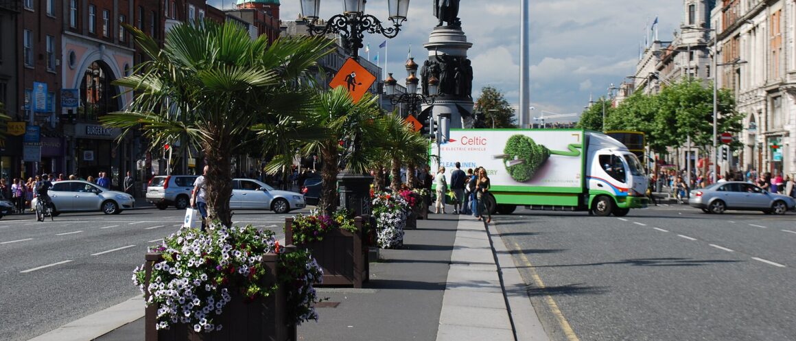 Flower planters and ornate lamps line O'Connell Bridge in Dublin, facing the O'Connell Monument and busy street.