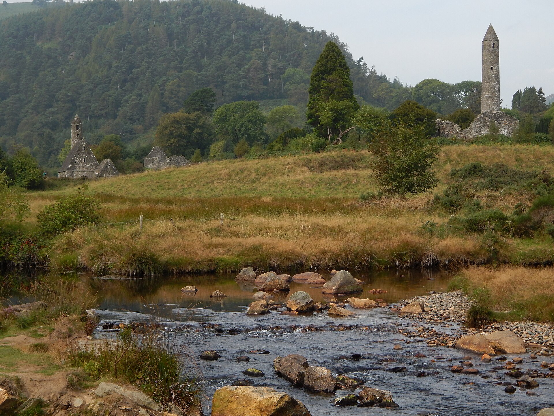 Glendalough round tower and stone church ruins stand across a rocky stream beneath a wooded hillside.