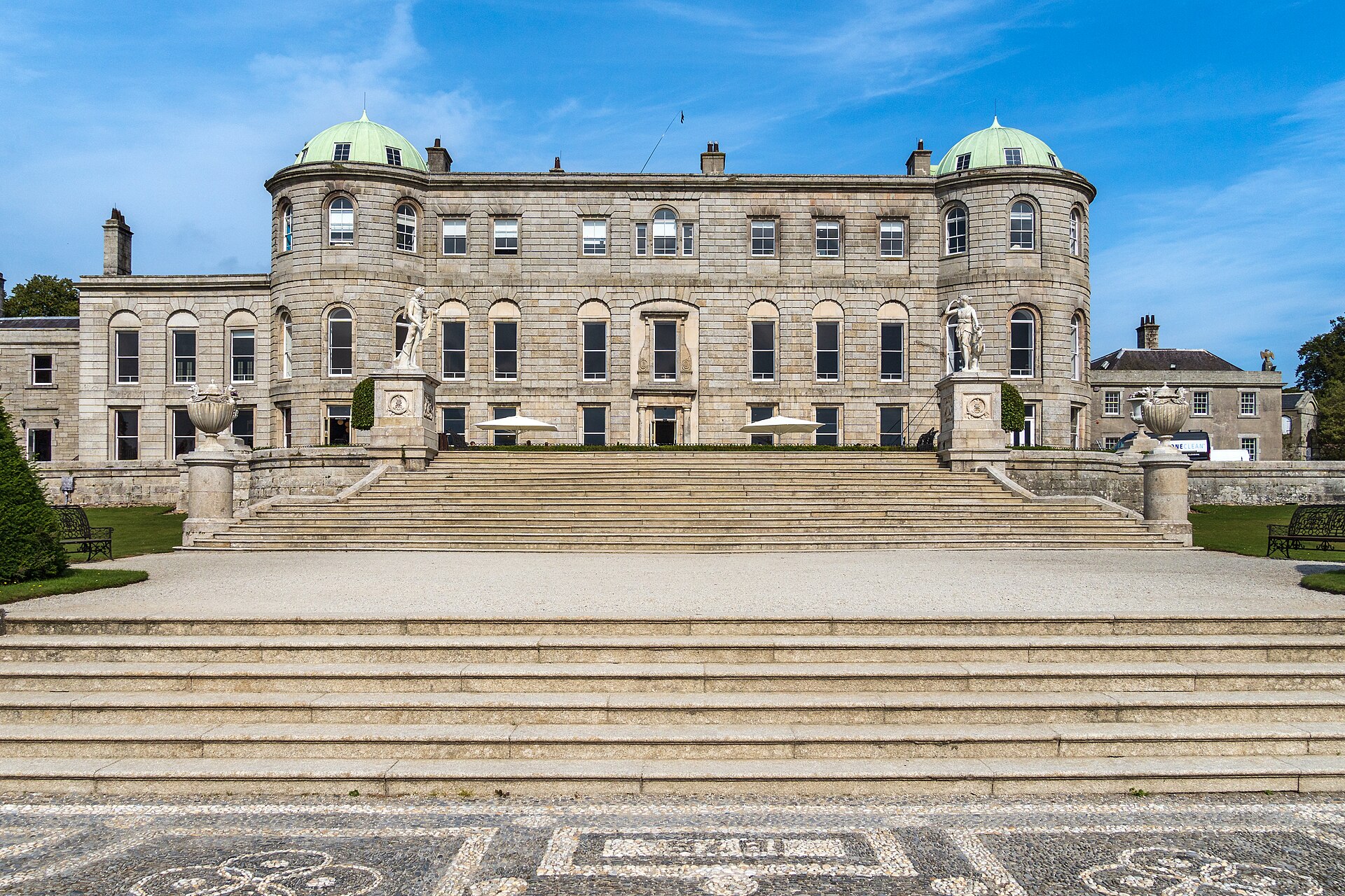 Granite garden front of Powerscourt House in Co. Wicklow, with two green domes, statues, broad steps and white parasols on the terrace under a blue sky.