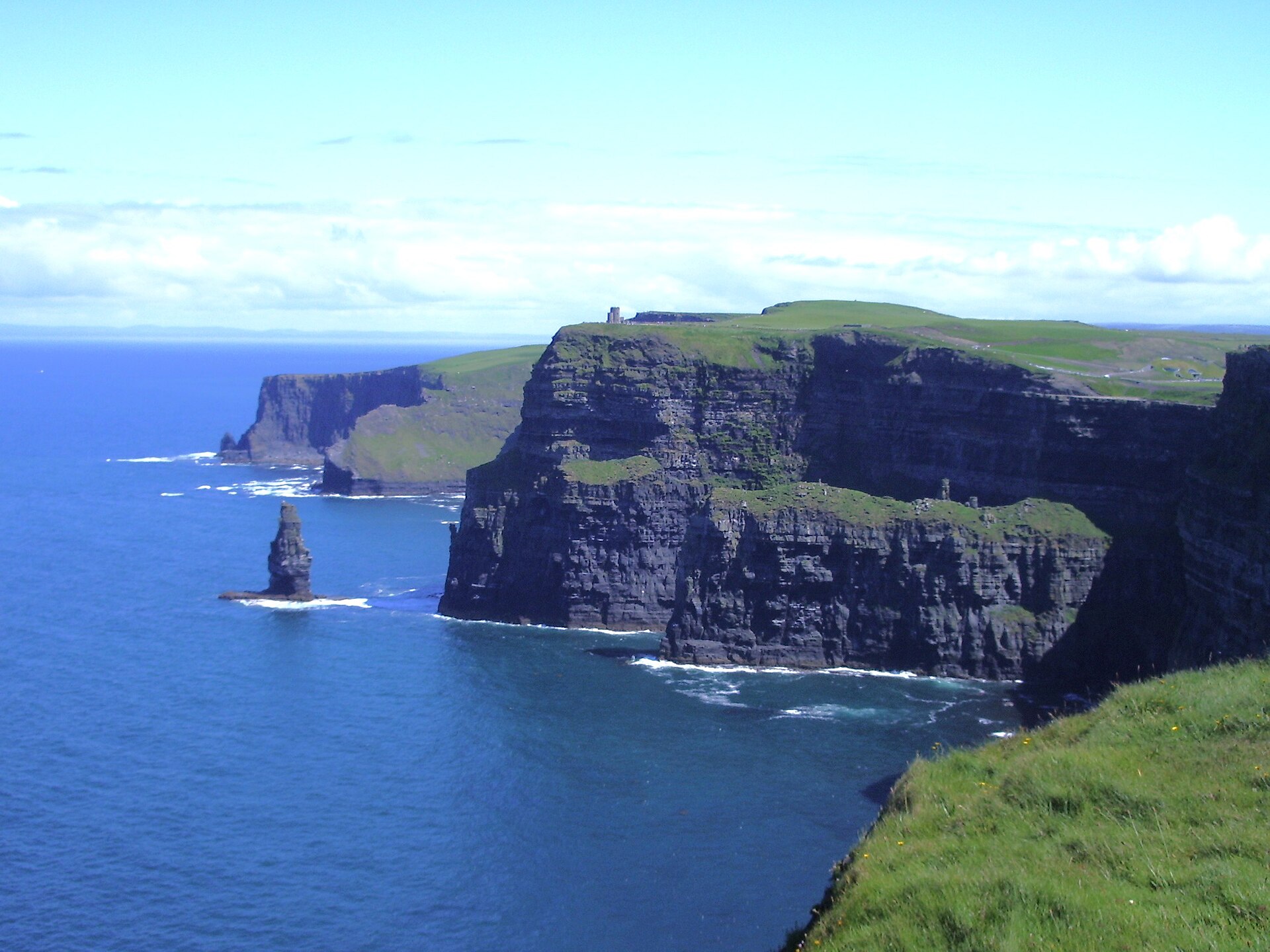 Grass-topped Cliffs of Moher rise above the Atlantic, with a narrow sea stack offshore and white surf below.
