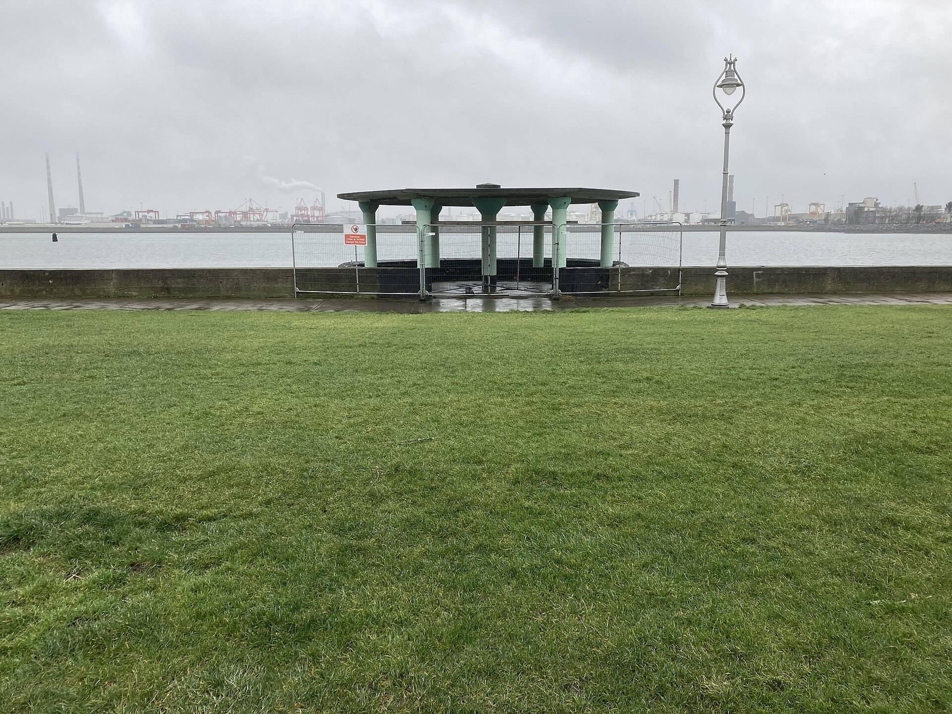 A green bandstand stands beside the seawall on Clontarf Promenade in Dublin, with grass and harbour views beyond.