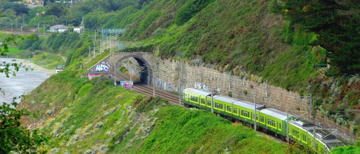 A green DART train follows the coastal railway between Killiney and Dalkey, beneath a wooded hillside beside a stone tunnel.