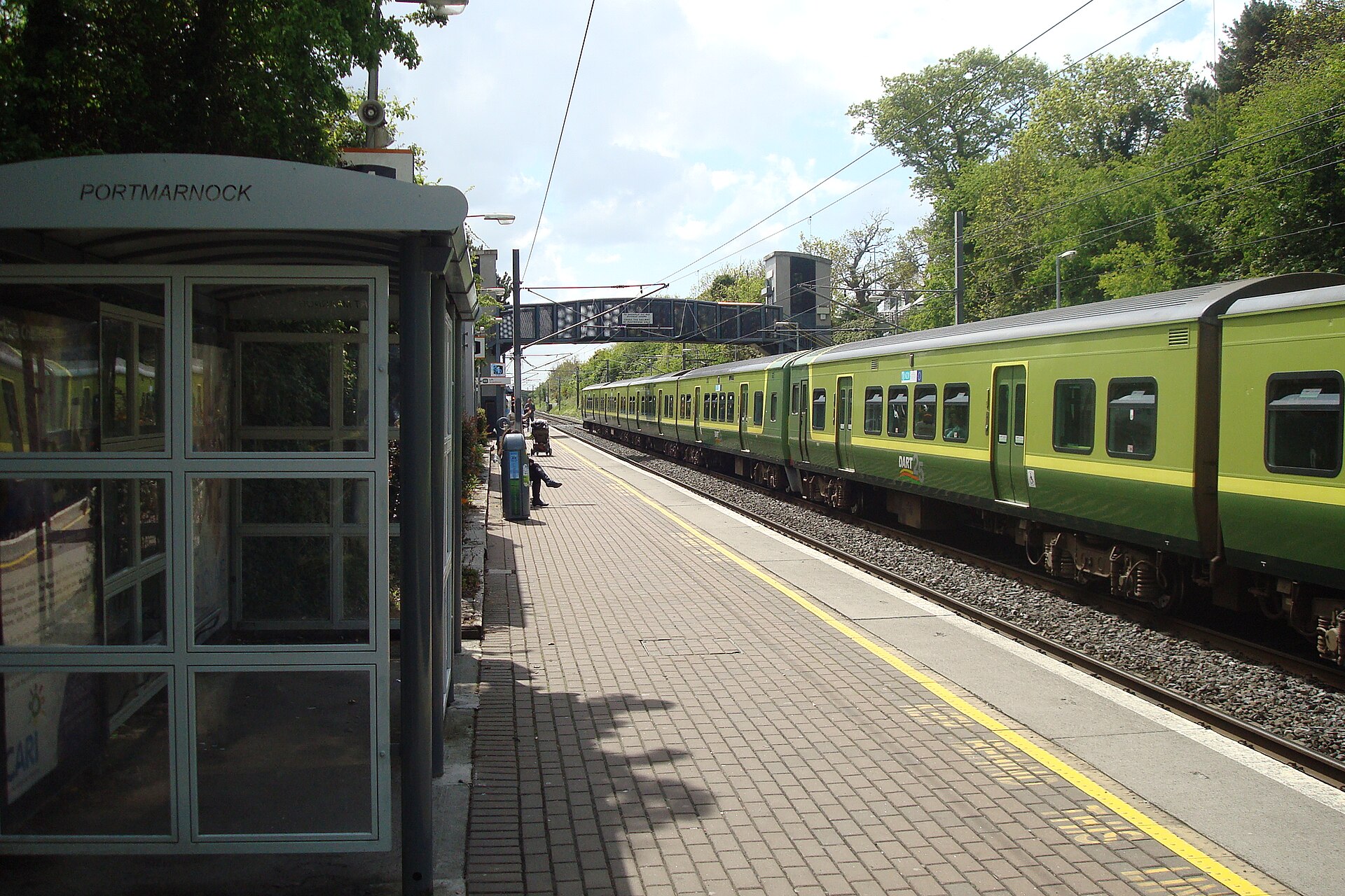 A green DART train runs beside the paved platform and shelter at Portmarnock station on Dublin's northern coastal line.