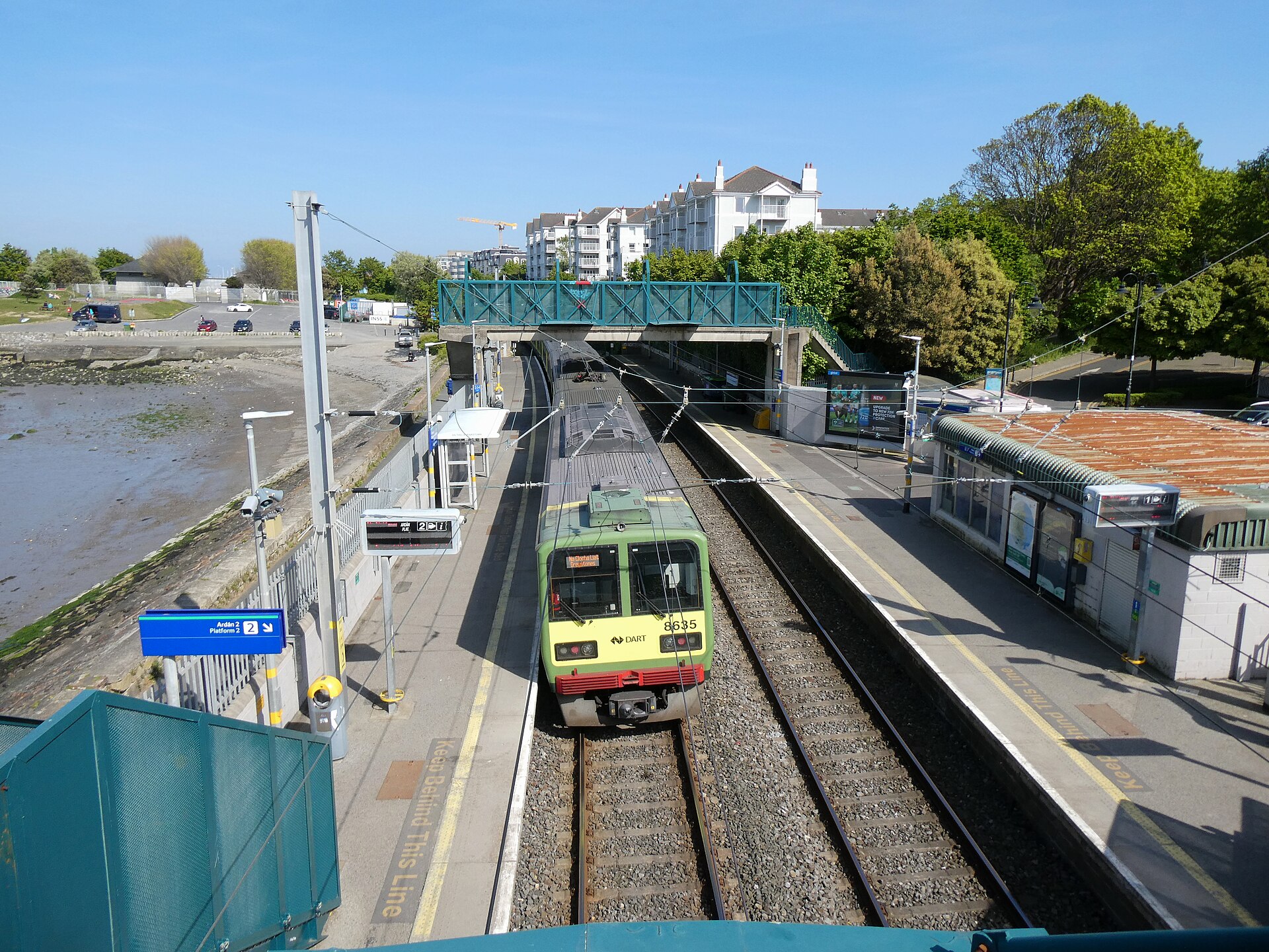 A green DART train stands between platforms at Salthill and Monkstown, with the tidal shore beside the station.