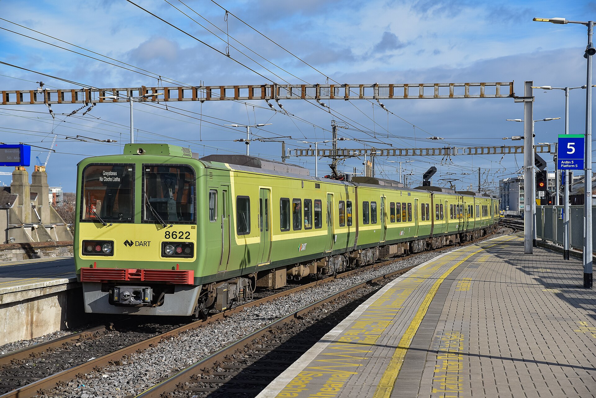 A green DART train stands beside a curved platform at Dublin Connolly station, beneath overhead wires and a blue sky.