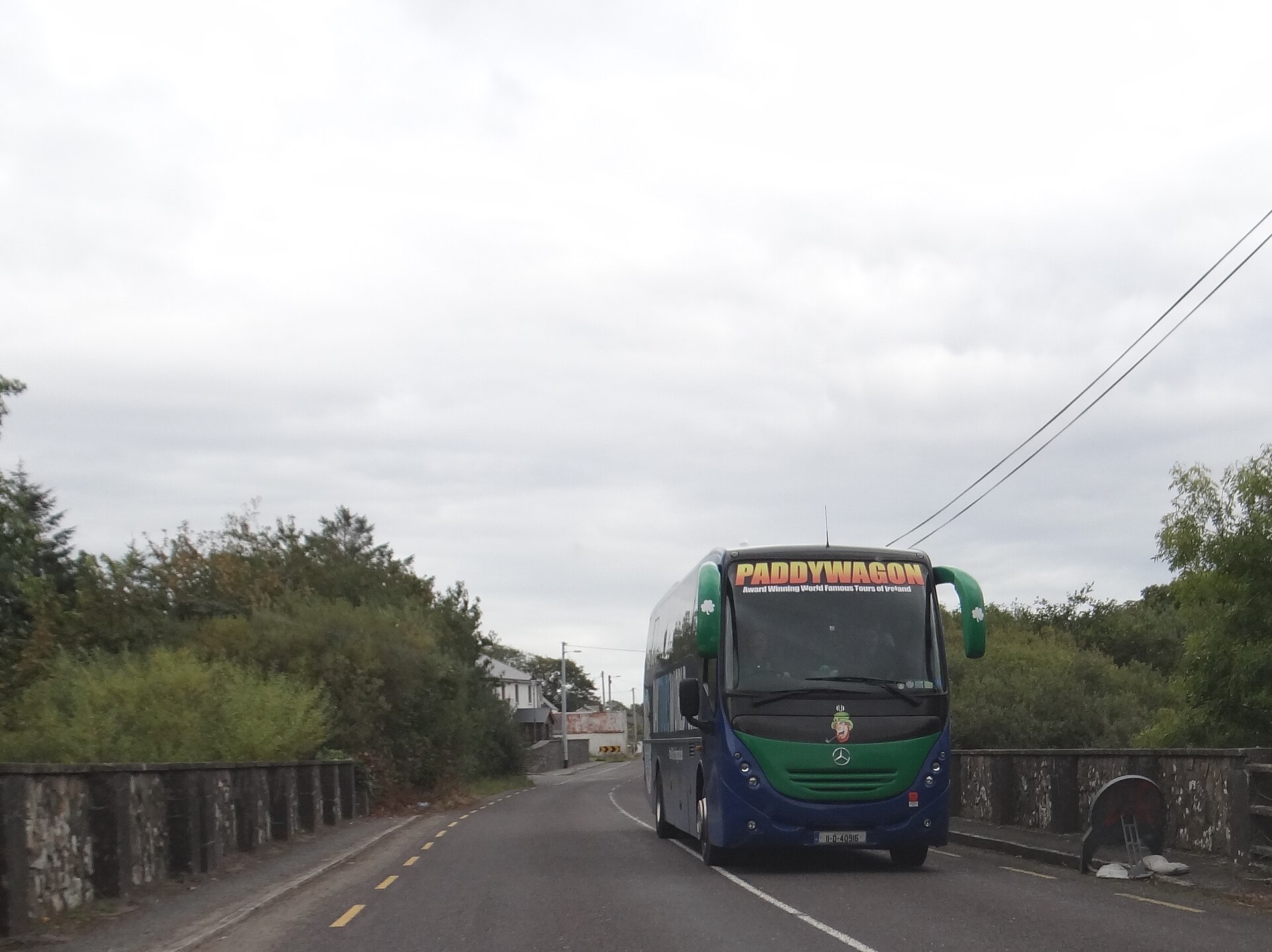 A green Paddywagon tour coach approaches along a narrow road between stone parapets and hedges in County Kerry, Ireland.