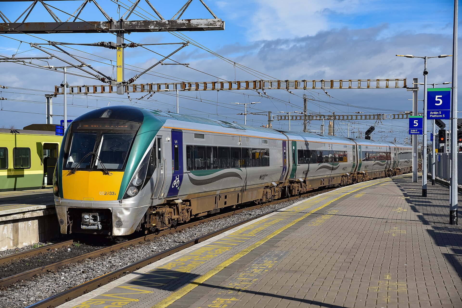 A green and silver Irish Rail railcar stands alongside platform five at Dublin Connolly in sunshine.
