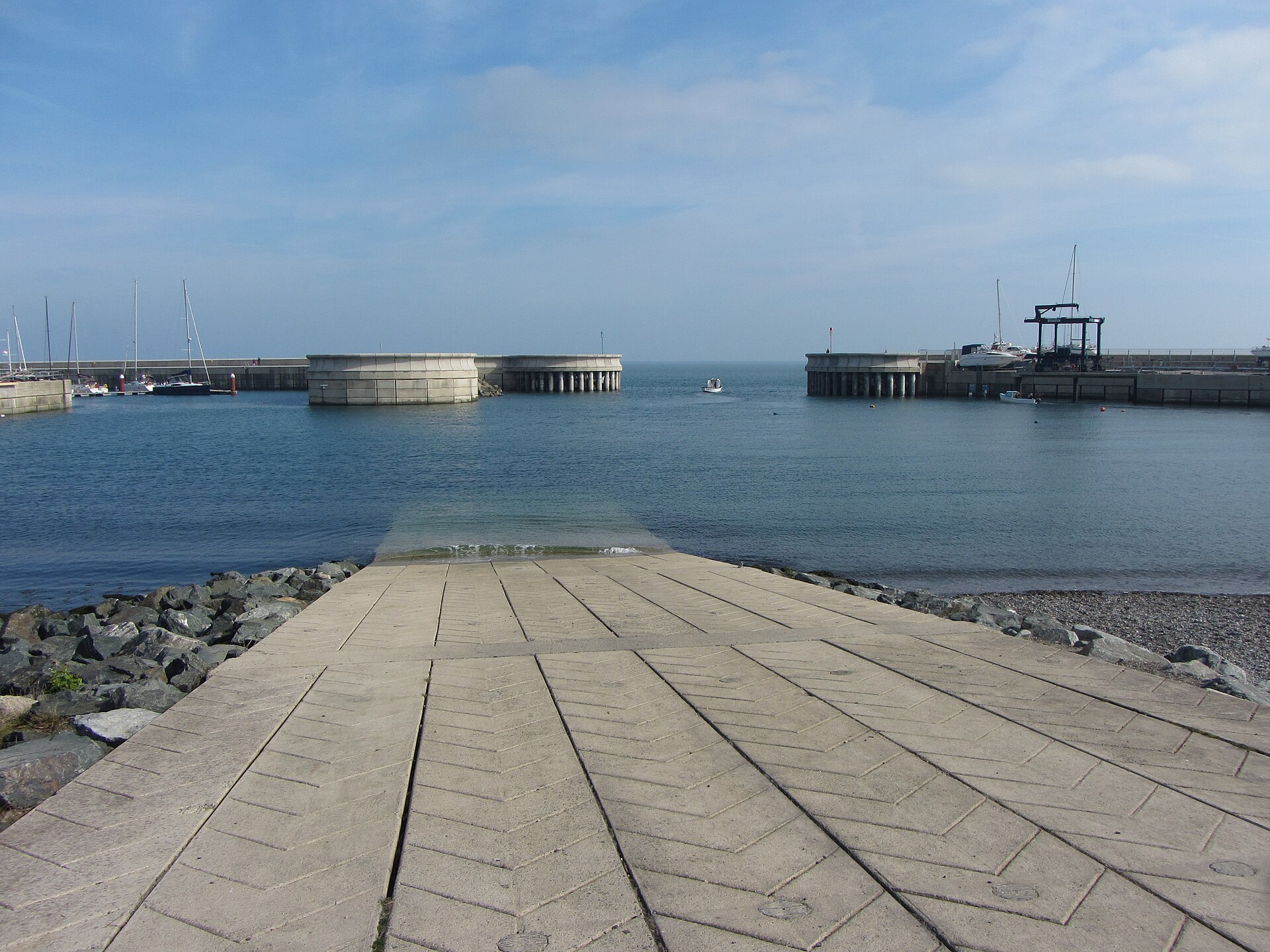 A grooved concrete slipway leads into Greystones Harbour, facing the harbour entrance with sailing boats to the left.