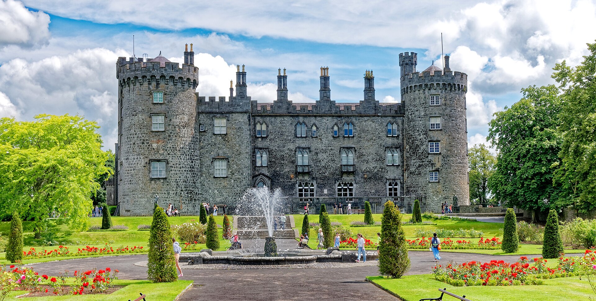 Kilkenny Castle's round towers rise behind a fountain, clipped conifers and red flower beds, with visitors walking through the garden.