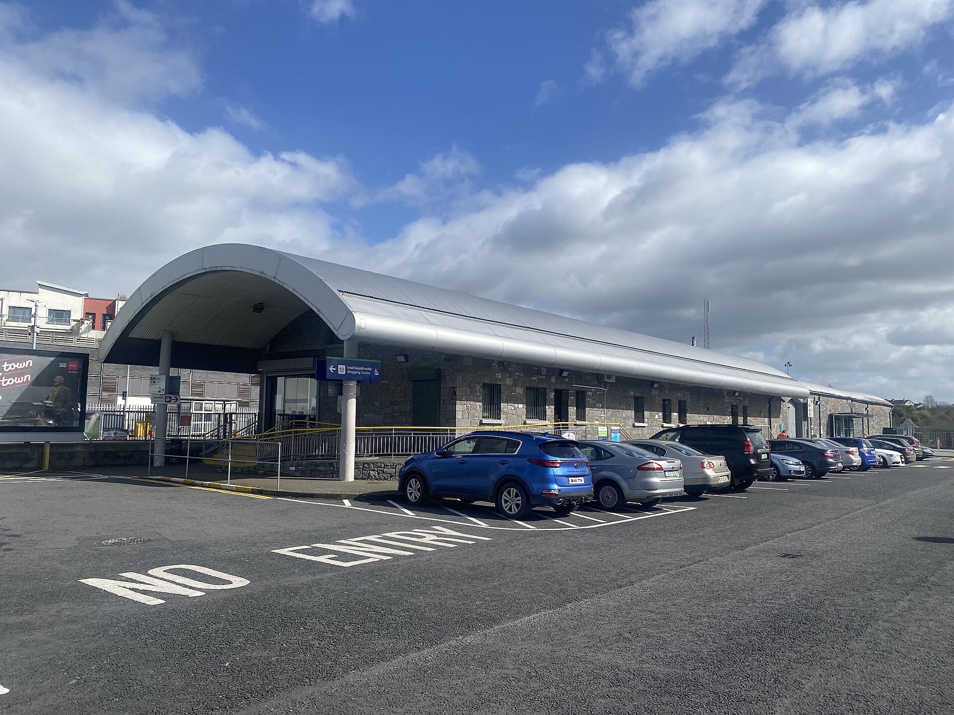 Kilkenny railway station's stone entrance building has a curved silver canopy, with a ramp and parked cars outside.