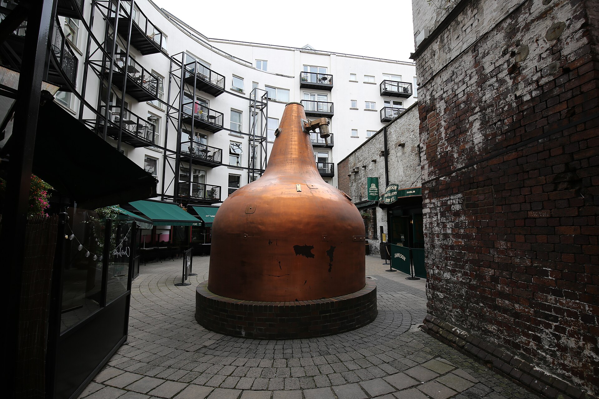A large copper pot still stands in the paved courtyard at Jameson Distillery Bow St, surrounded by brickwork and balconies.