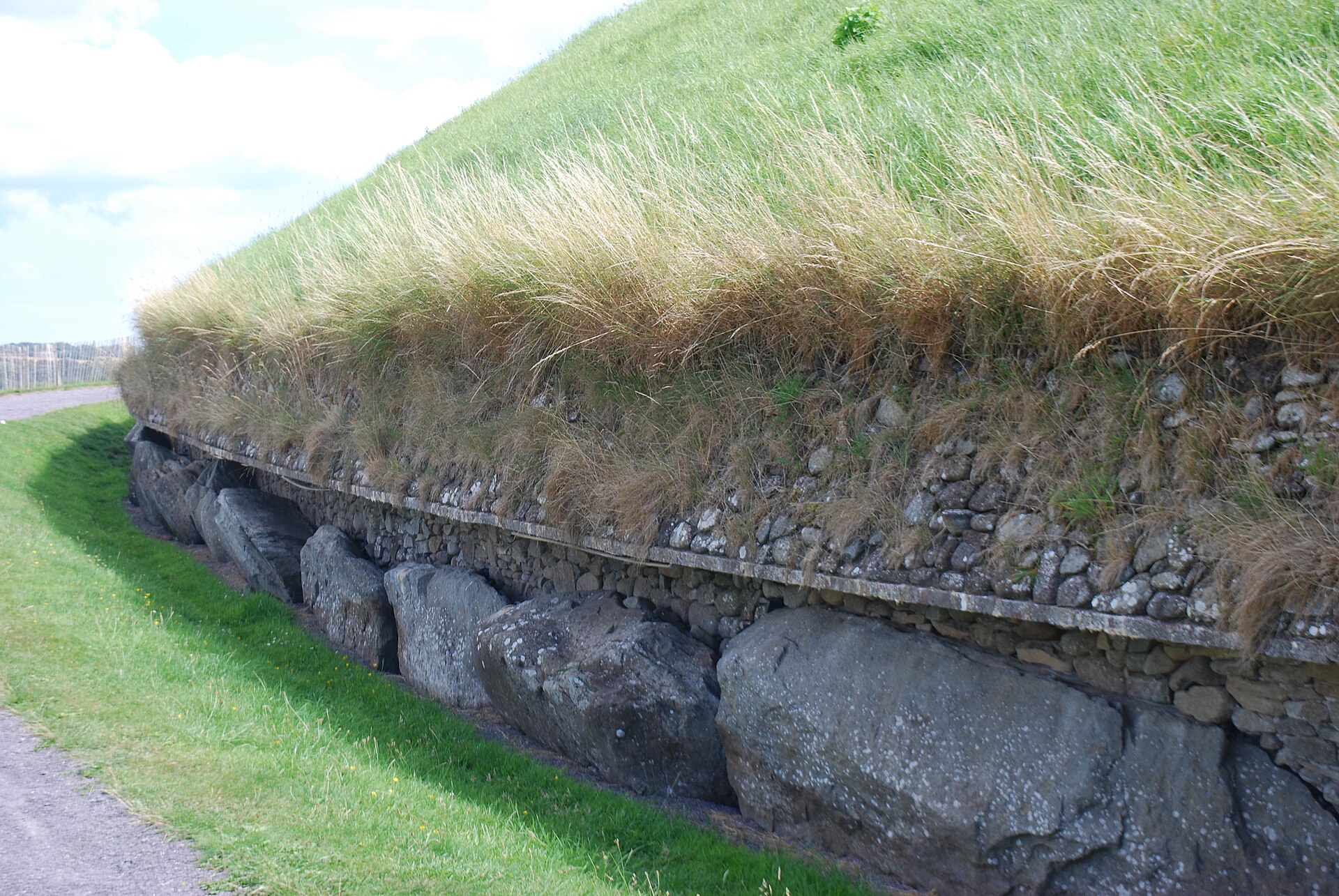 Large kerbstones curve around the base of Newgrange, below rough stonework and the overhanging grass of the mound.