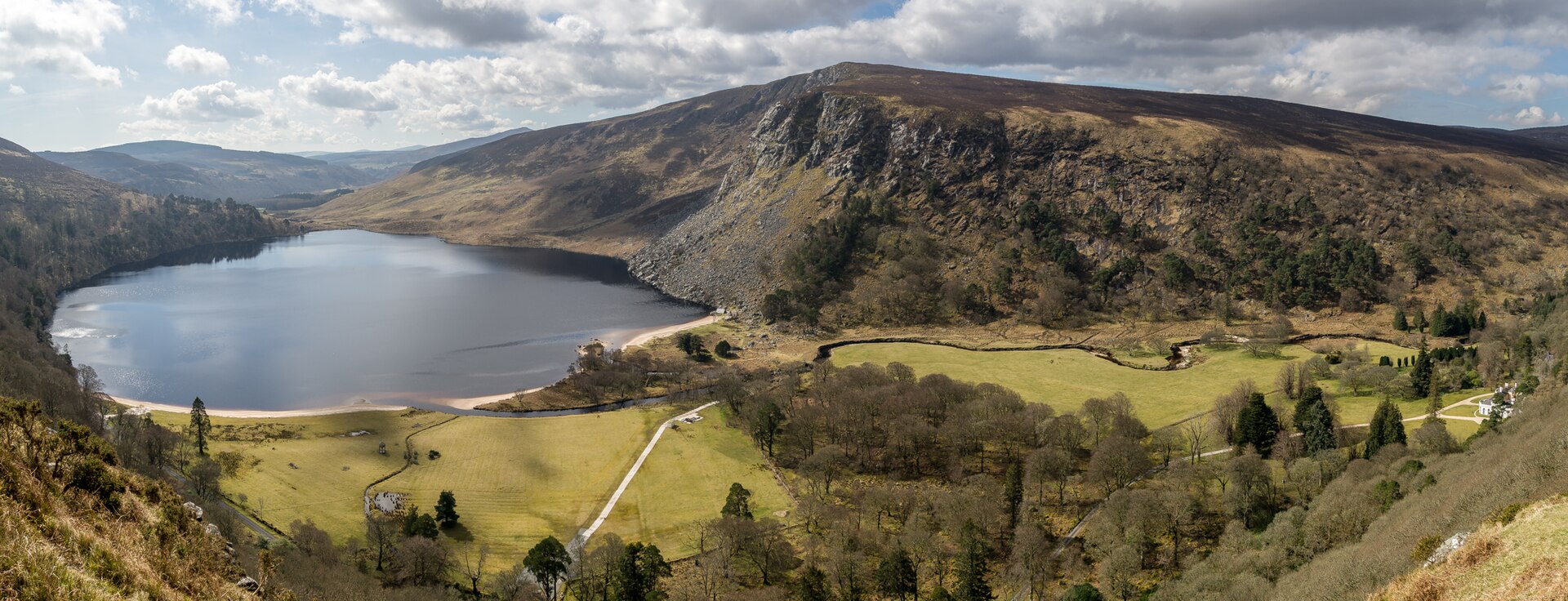 Lough Tay lies beneath Luggala's steep rocky slopes, bordered by a pale beach, fields and woodland in Wicklow.