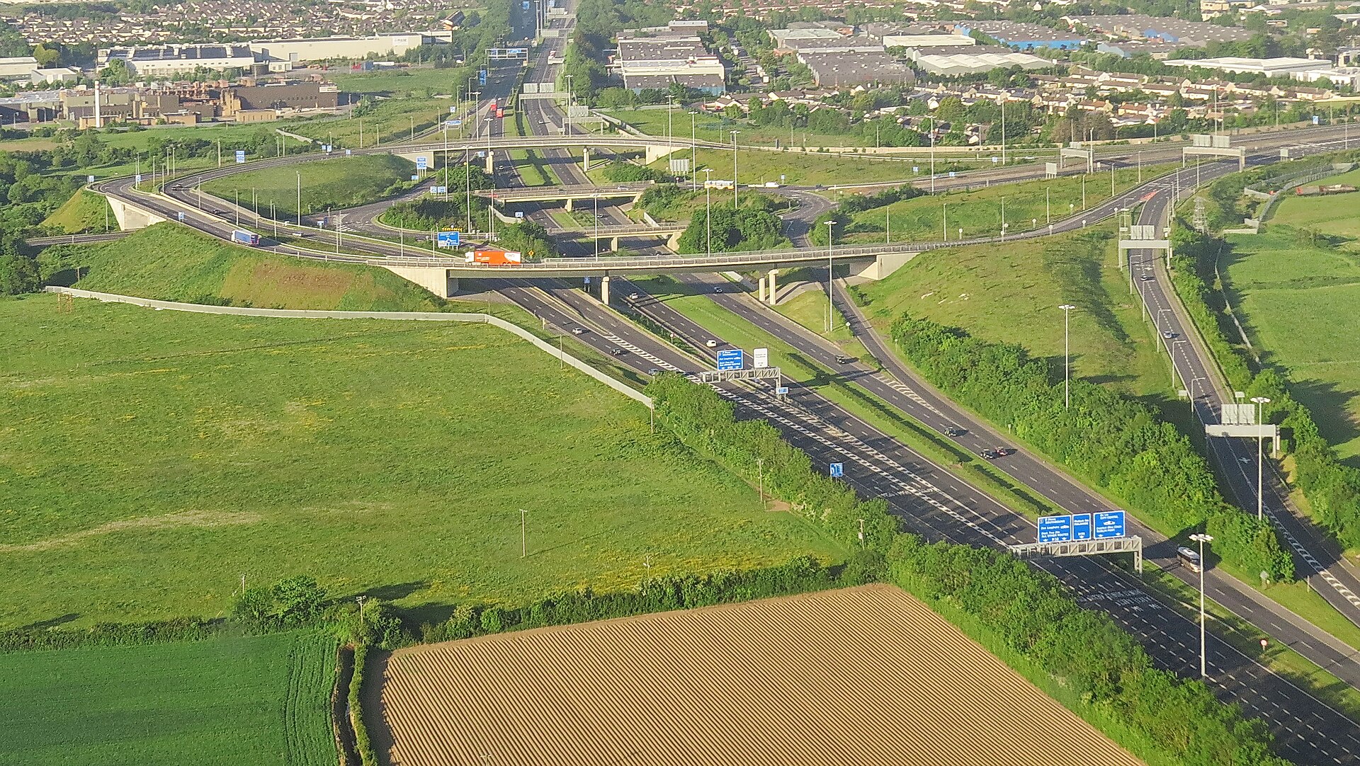 The M1 motorway at Swords crosses green fields beneath bridges, with slip roads and blue overhead direction signs.