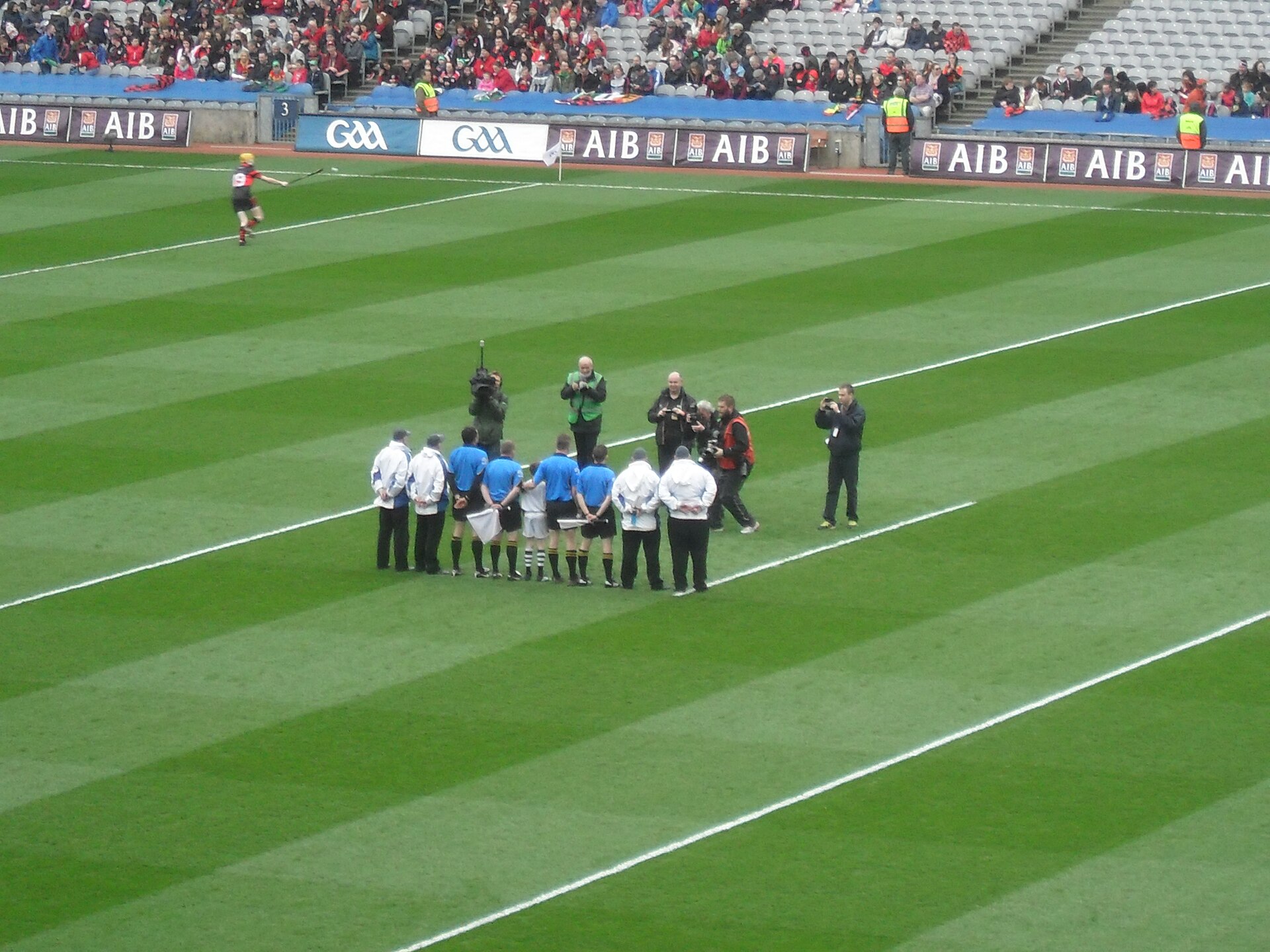 Match officials and photographers gather on the Croke Park pitch in Dublin, with spectators seated behind them.