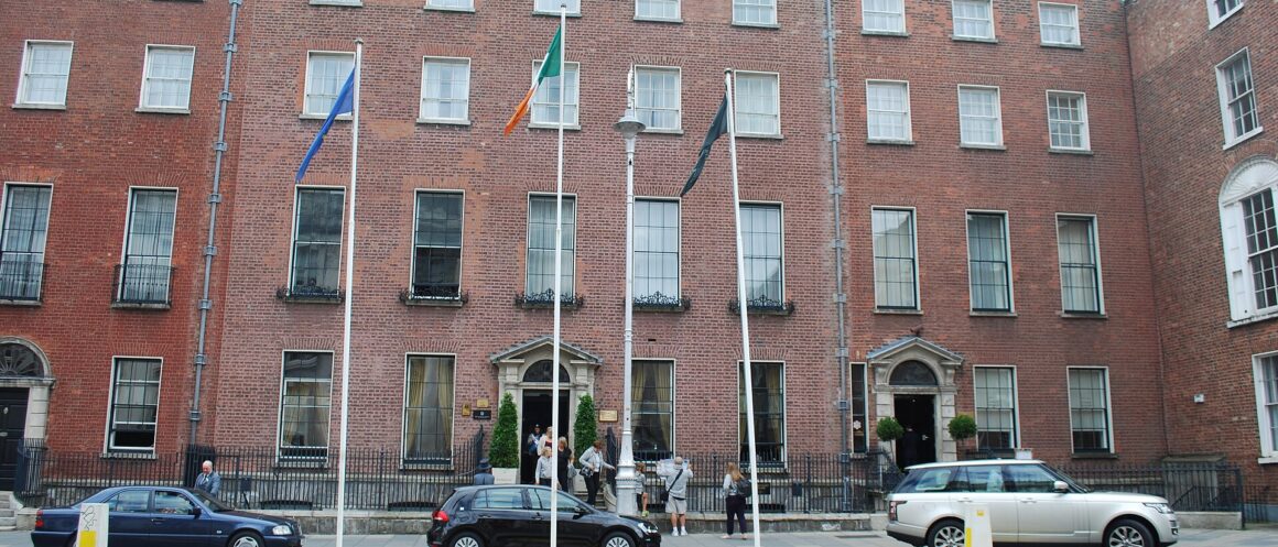 The Merrion Hotel in Dublin has a red brick Georgian frontage, tall sash windows, flagpoles and pedestrians by its doorways.