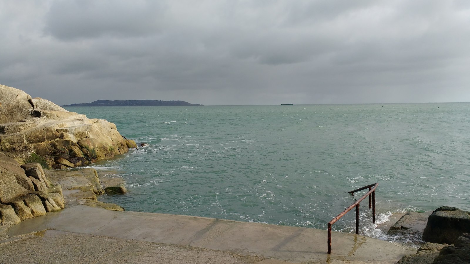 A metal handrail descends from the concrete bathing platform into the sea at the Forty Foot, beside large coastal rocks.