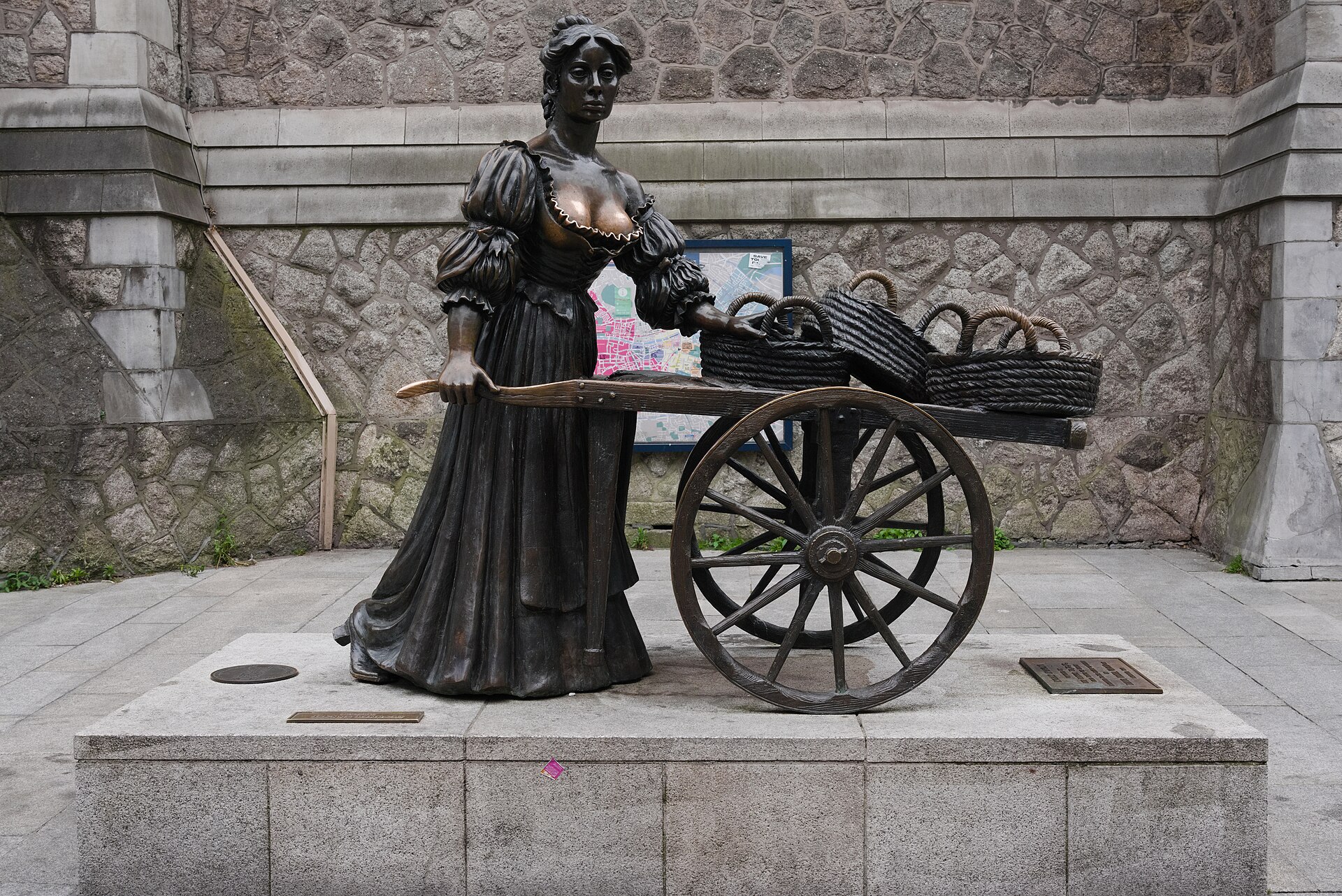 Molly Malone's bronze statue and basket-filled handcart stand against a stone wall on Suffolk Street, Dublin.