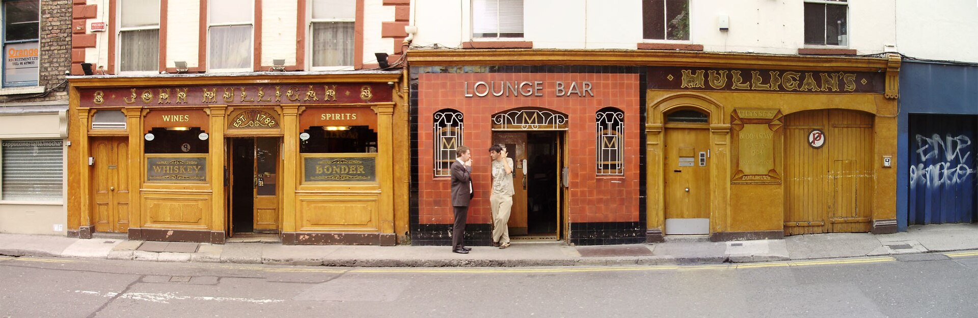 Mulligan's in Dublin, with ochre wooden doors and windows flanking a red tiled lounge entrance where two people stand.