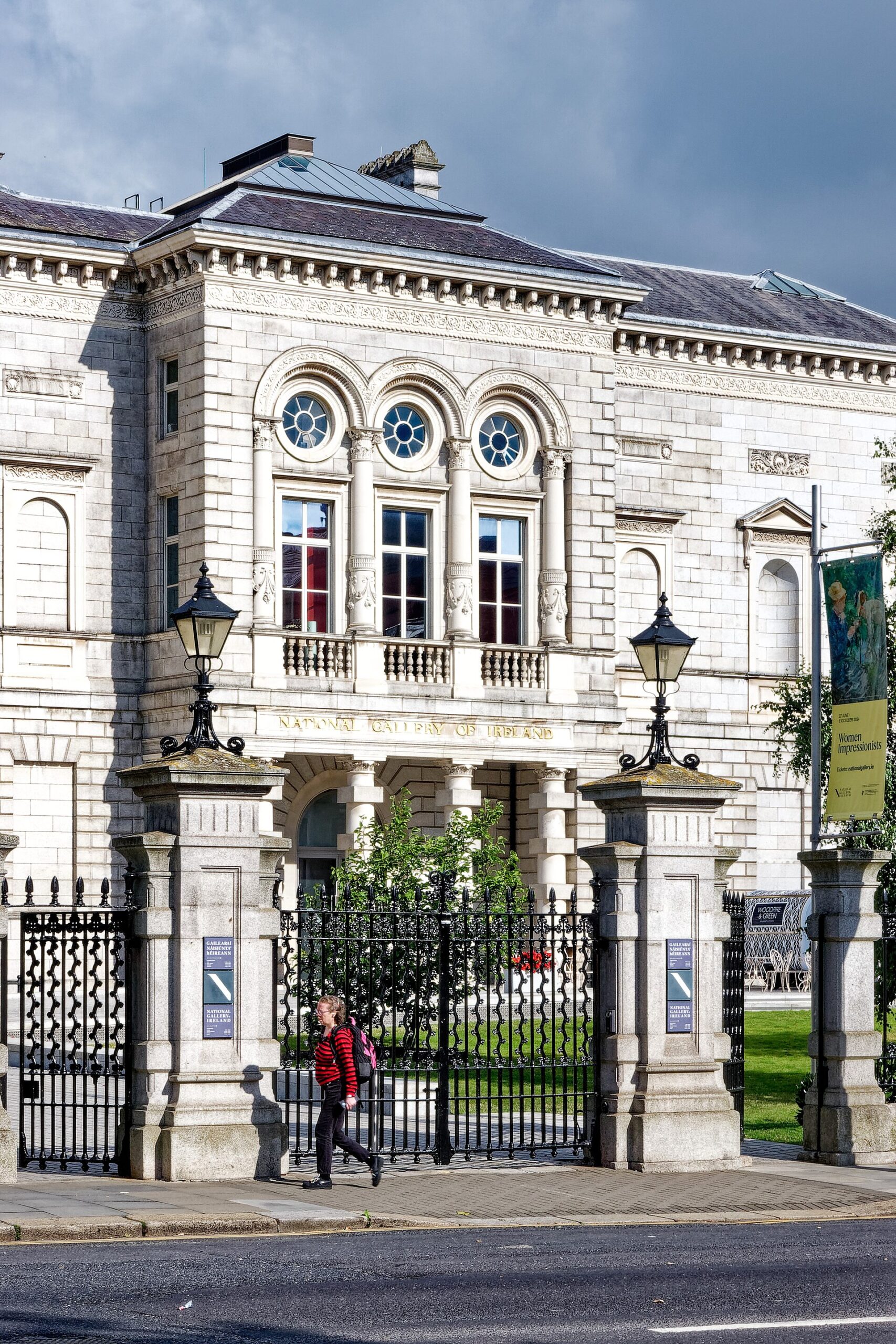 The National Gallery of Ireland facade in Dublin, with arched windows, iron gates and a pedestrian outside.