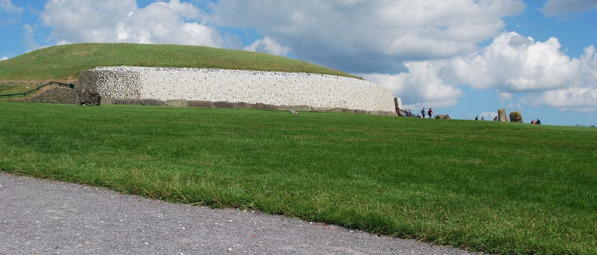 Newgrange passage tomb rises above a green lawn, its white quartz facade beneath a grassy mound and blue sky.