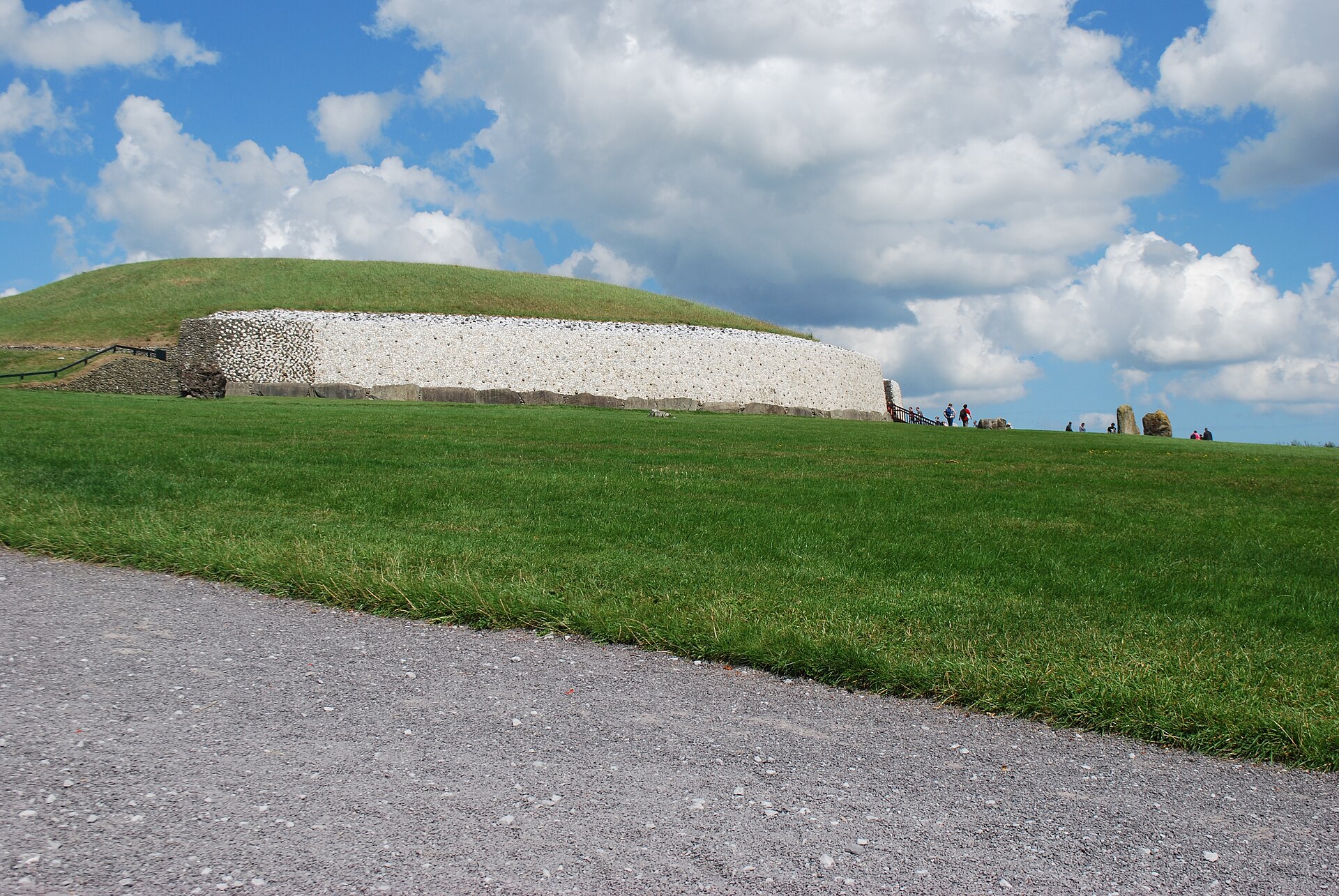 Newgrange passage tomb rises above a green lawn, its white quartz facade beneath a grassy mound and blue sky.