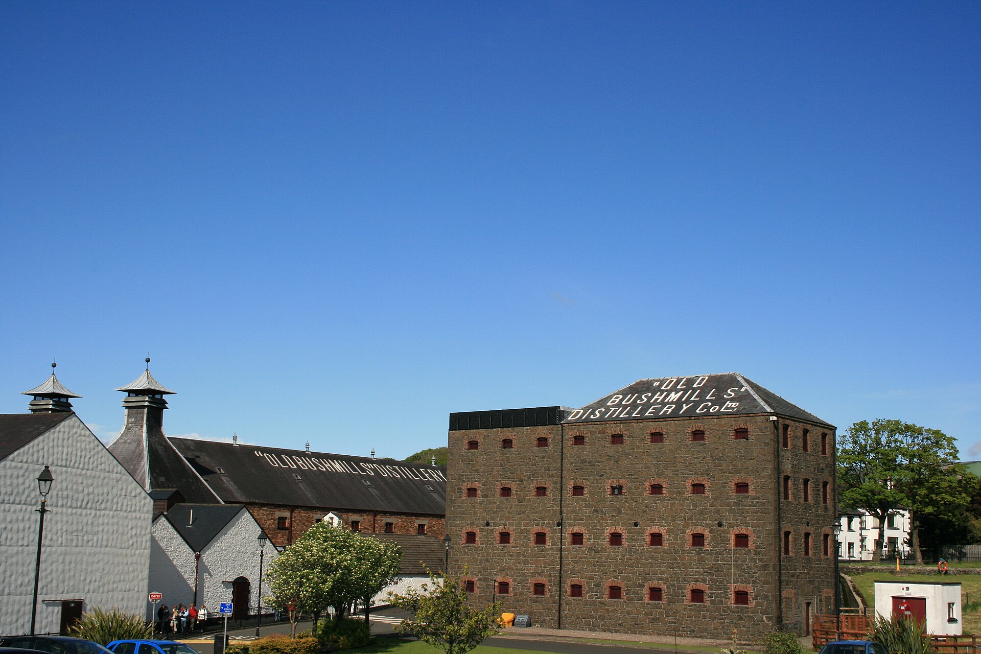 Old Bushmills Distillery's stone warehouse and white buildings stand beneath a blue sky, with the name painted across the roofs.