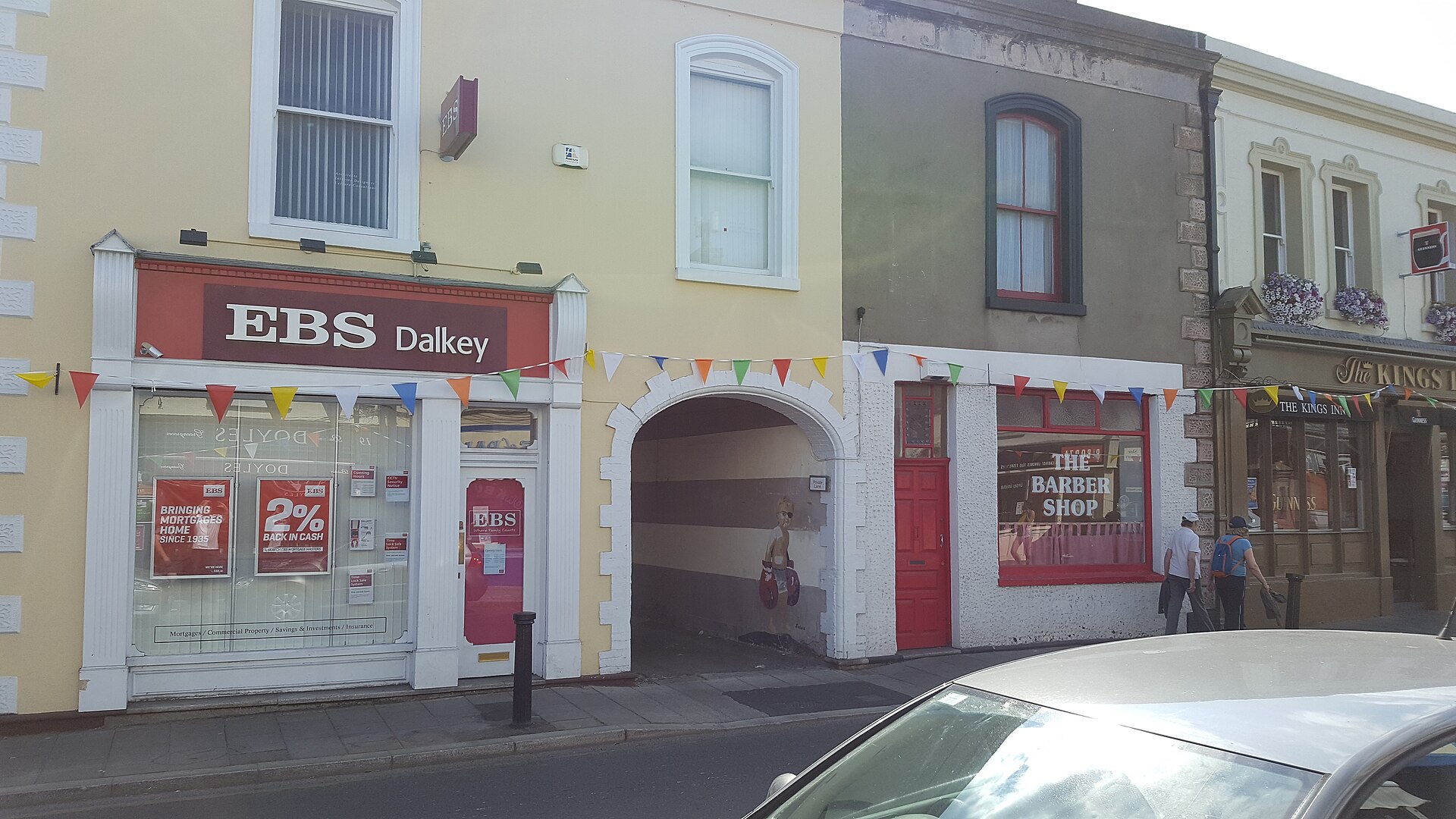Painted shopfronts and an arched passage line Castle Street in Dalkey village, with bunting overhead and pedestrians on the pavement.