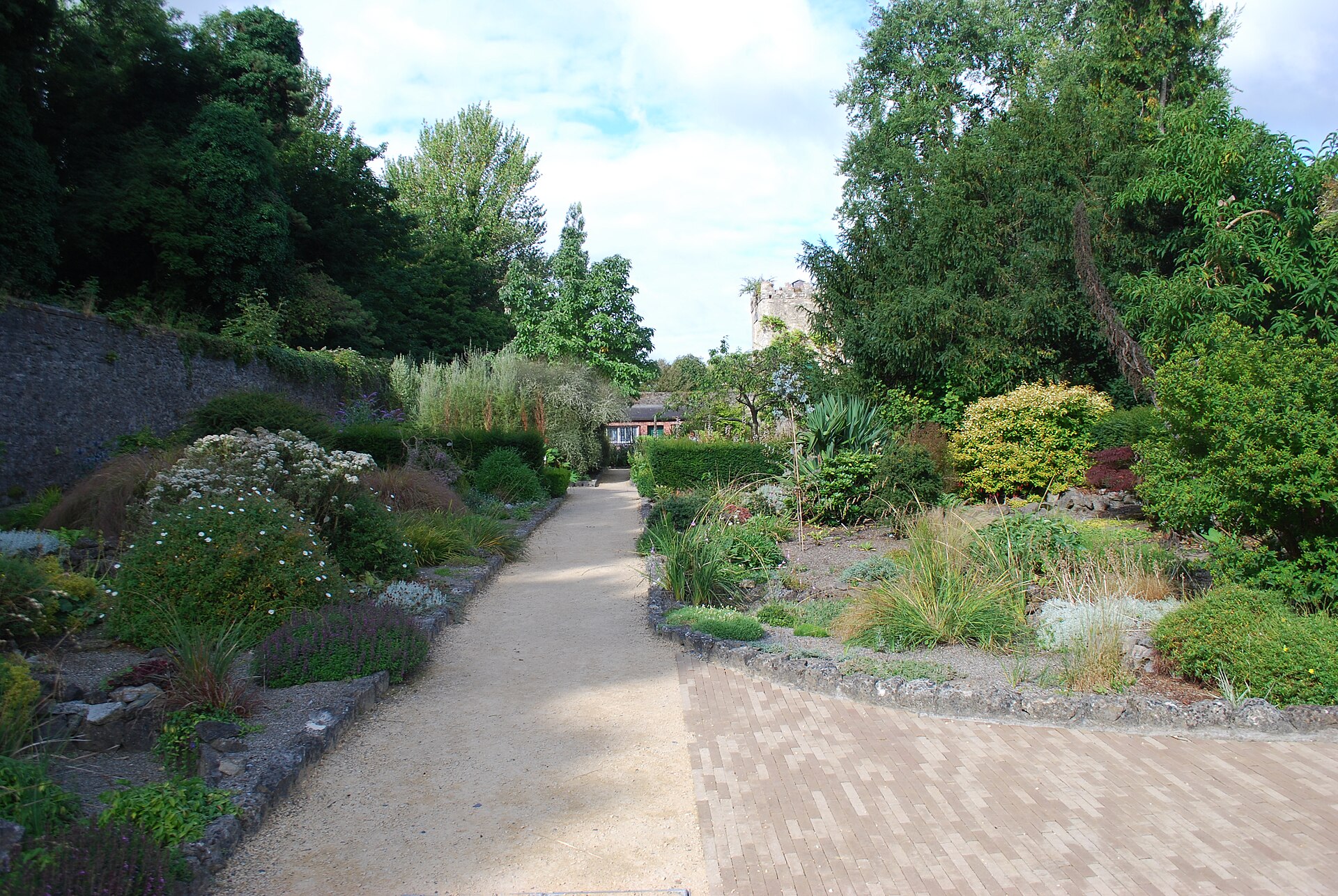 A pale path winds between planted borders, mature trees and a stone wall in the gardens at Malahide Castle.