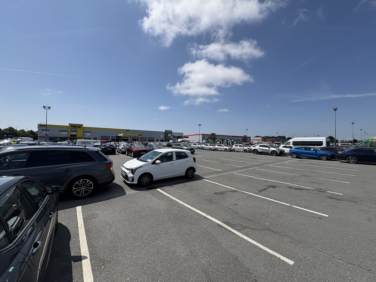 Parked cars fill the Dublin Airport rental area, with hire-company buildings beyond the marked parking bays.