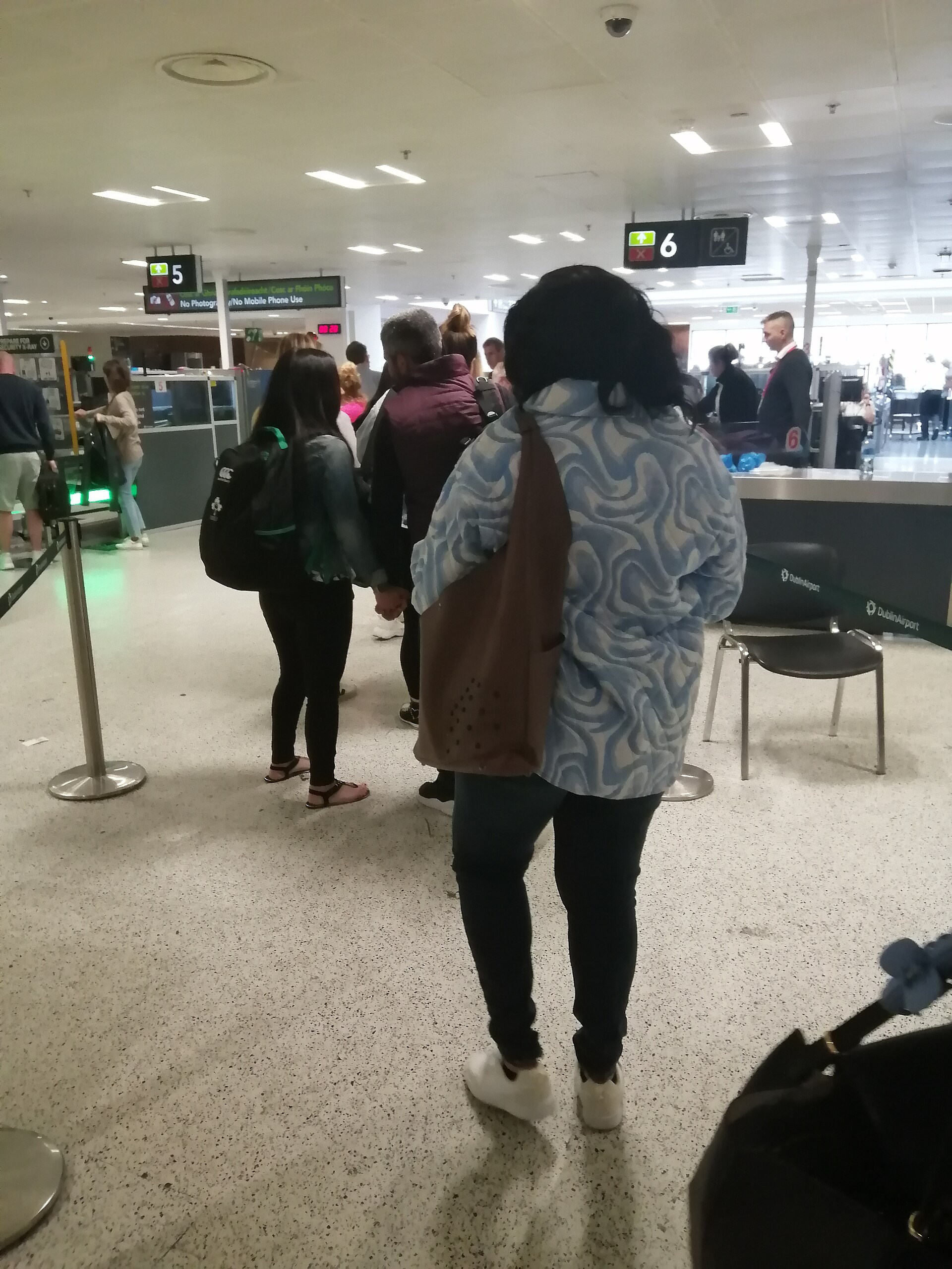 Passengers queue at Dublin Airport security screening lanes, with baggage trays and screening equipment ahead.