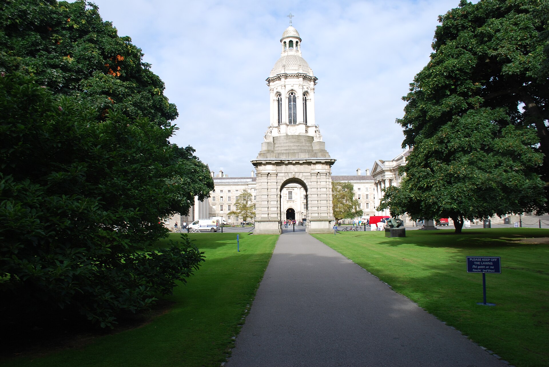 A path between lawns leads to Trinity College Dublin's stone Campanile, framed by mature trees and college buildings.