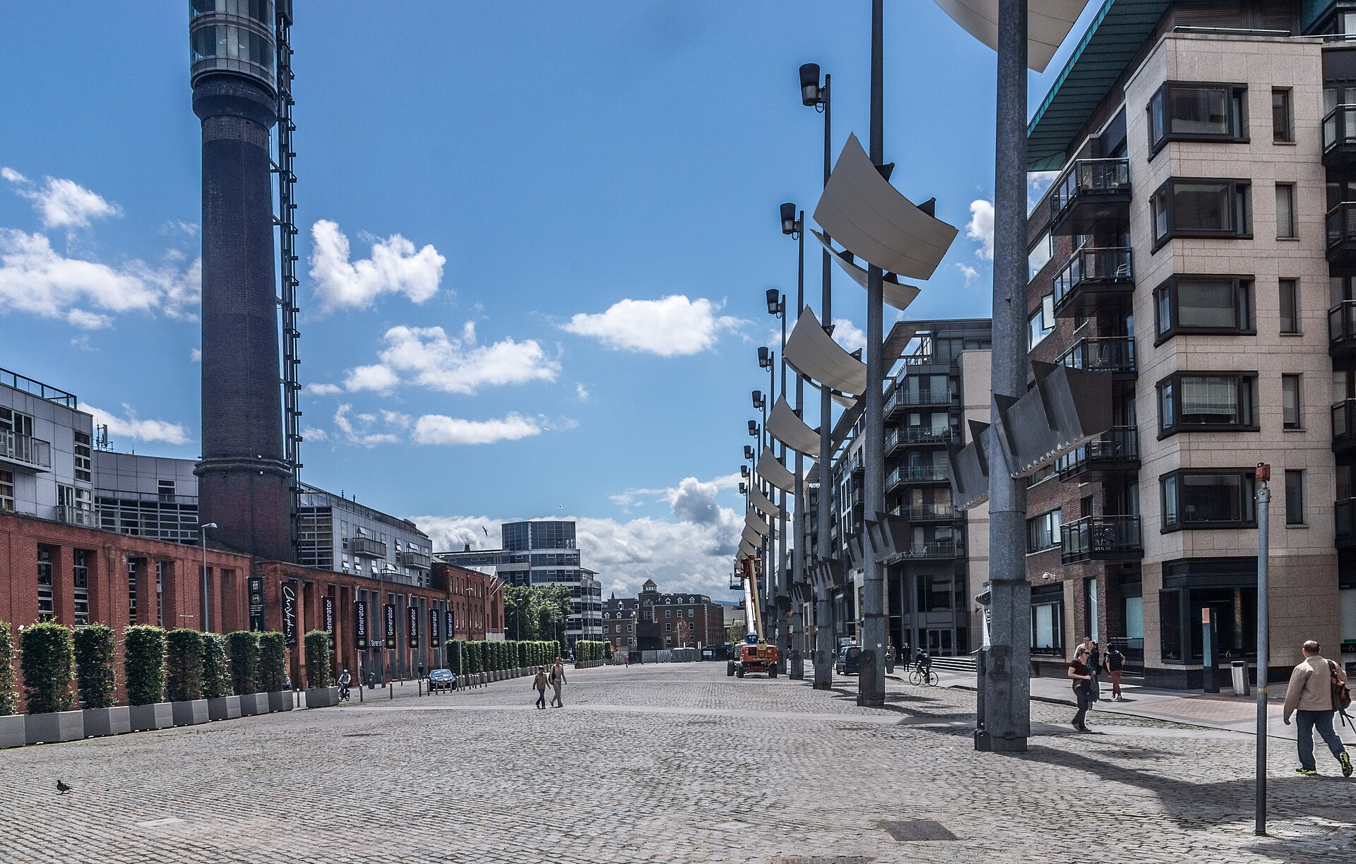 Pedestrians cross the broad cobbled Smithfield Square in Dublin, between the distillery chimney and a row of modern apartment buildings.